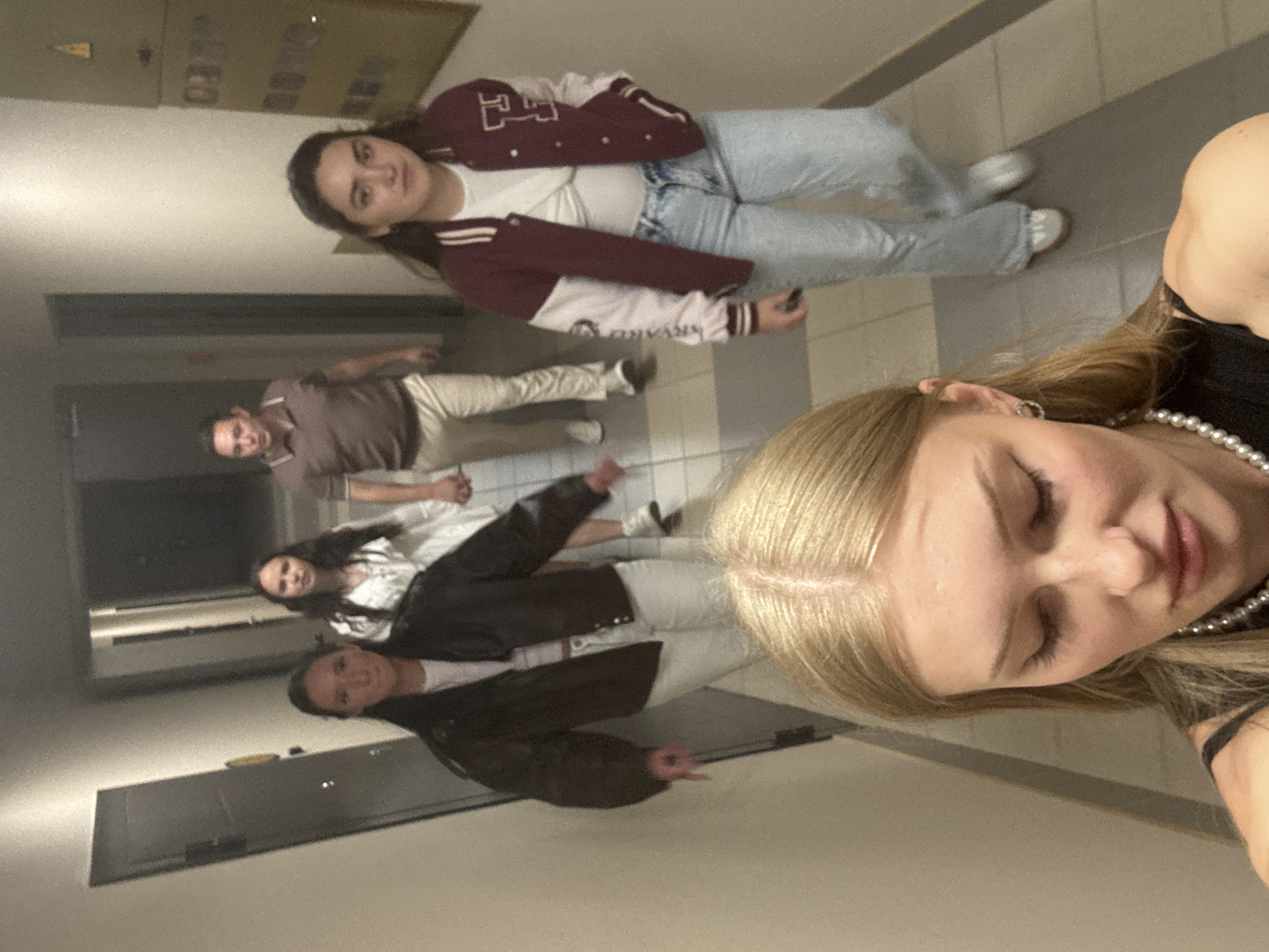 The image shows five individuals in a dimly lit hallway, likely in a residential building or apartment complex in the city of Perm, Russia. In the foreground, a young woman with blonde hair and a pearl necklace is looking down, her face partially visible. Behind her, walking away from the camera, are four other people.
The person immediately behind the blonde woman, on the left, is wearing a dark bomber jacket and white pants. To their right and slightly further back, a woman in a white dress is walking. Further back still, on the right side of the hallway, a woman is wearing a burgundy and white bomber jacket and light-wash jeans. Behind her, a man in a brown polo shirt and light-colored pants is walking.
The hallway has beige walls and a tiled floor. A dark door is visible on the left side of the image. A metal box with various compartments is mounted on the wall on the right side. The lighting suggests it is evening or nighttime. The overall impression is of a candid moment captured as a group walks down a hallway.