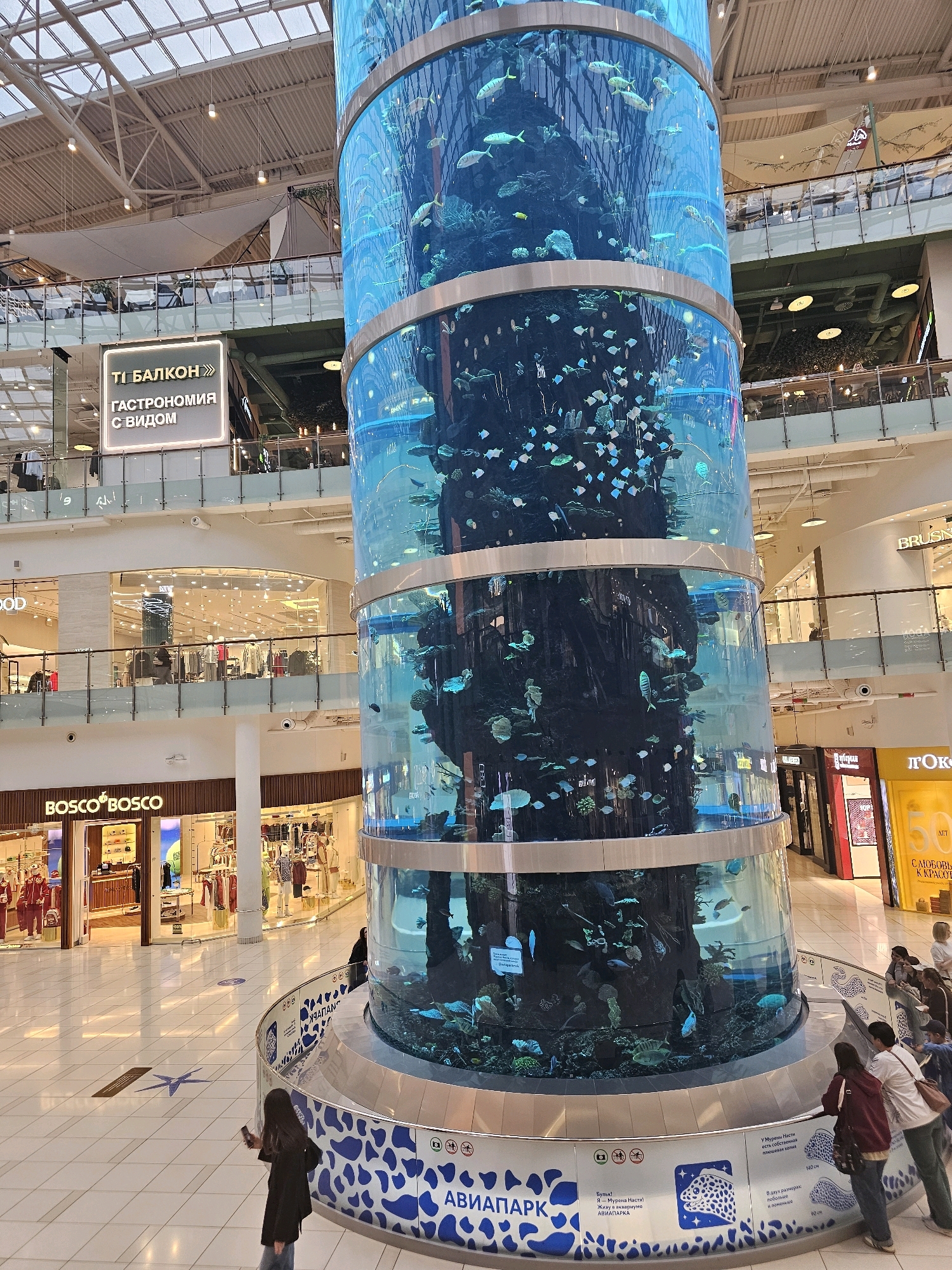 The image shows a large, cylindrical aquarium in the center of a multi-level shopping mall. The aquarium is filled with water and contains numerous fish and marine life. The mall has a modern design with shops, escalators, and glass railings visible. Several people are present, observing the aquarium or shopping. One person is standing in the foreground, looking at the aquarium, while others are scattered throughout the mall on different levels. The lighting suggests it is daytime, with natural light filtering in from the ceiling and artificial lights illuminating the shops. The overall scene depicts a busy commercial environment with a prominent aquatic attraction.
