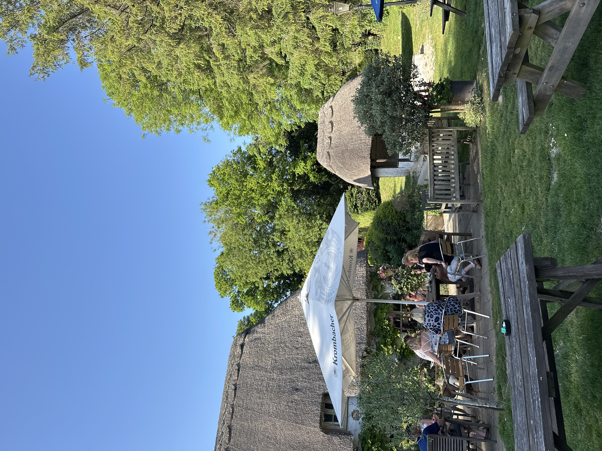 Here is a description of the image:
The image shows an outdoor seating area, possibly of a pub or restaurant, with a thatched-roof building in the background. People are seated at tables under a large white umbrella with the "Krombacher" logo. There are several wooden picnic tables on the grass and a small thatched-roof structure in the background, possibly a bus stop. The sky is blue and clear, with some trees visible above the buildings. The location is likely in a rural area with traditional architecture.