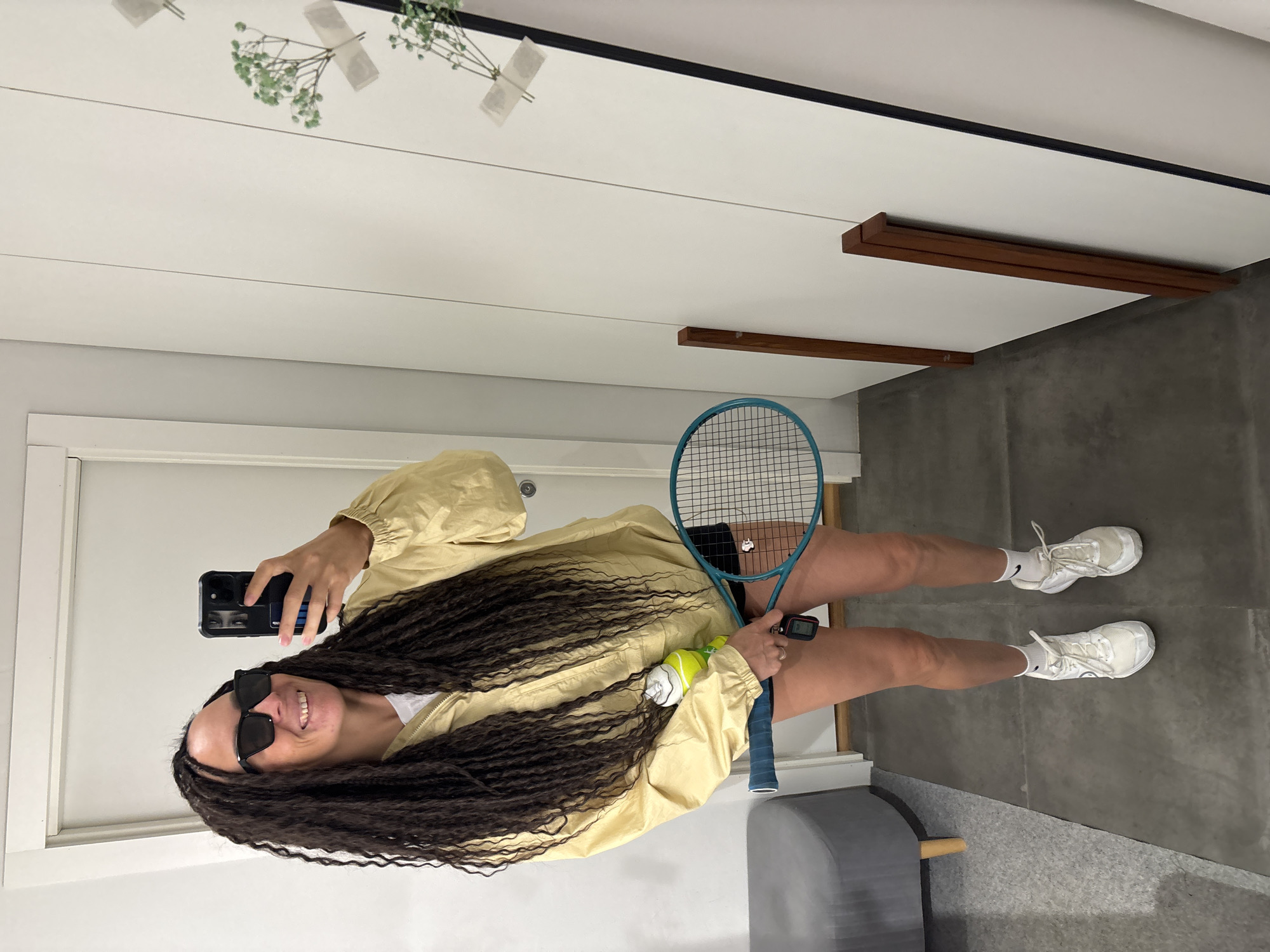 The image displays a full-body mirror selfie of an adult individual, likely female, captured indoors in what appears to be a modern setting in Chaikovsky, Russia. The subject has long, dark, braided hair, is wearing black sunglasses, and is smiling at the camera (her smartphone). She is dressed in a light yellow/beige long-sleeved jacket, black shorts, white socks, and white sneakers. She holds a black-cased smartphone in her left hand to take the photo, and in her right hand, she holds a blue-framed tennis racket along with two yellow-green tennis balls. A dark-colored device, possibly a fitness tracker or smartwatch, is visible on her left wrist.
The indoor scene features a large mirror against a white or light grey wall, reflecting parts of the room. The floor is dark grey, possibly concrete or large tiles. Reflected above the subject are small green plant sprigs, seemingly taped to the mirror, and two dark wooden shelves or beams mounted on a light-colored wall. A grey padded bench or stool is partially visible at the bottom left. The overall mood is casual and happy, suggesting an activity related to tennis. There is no discernible text in the image.