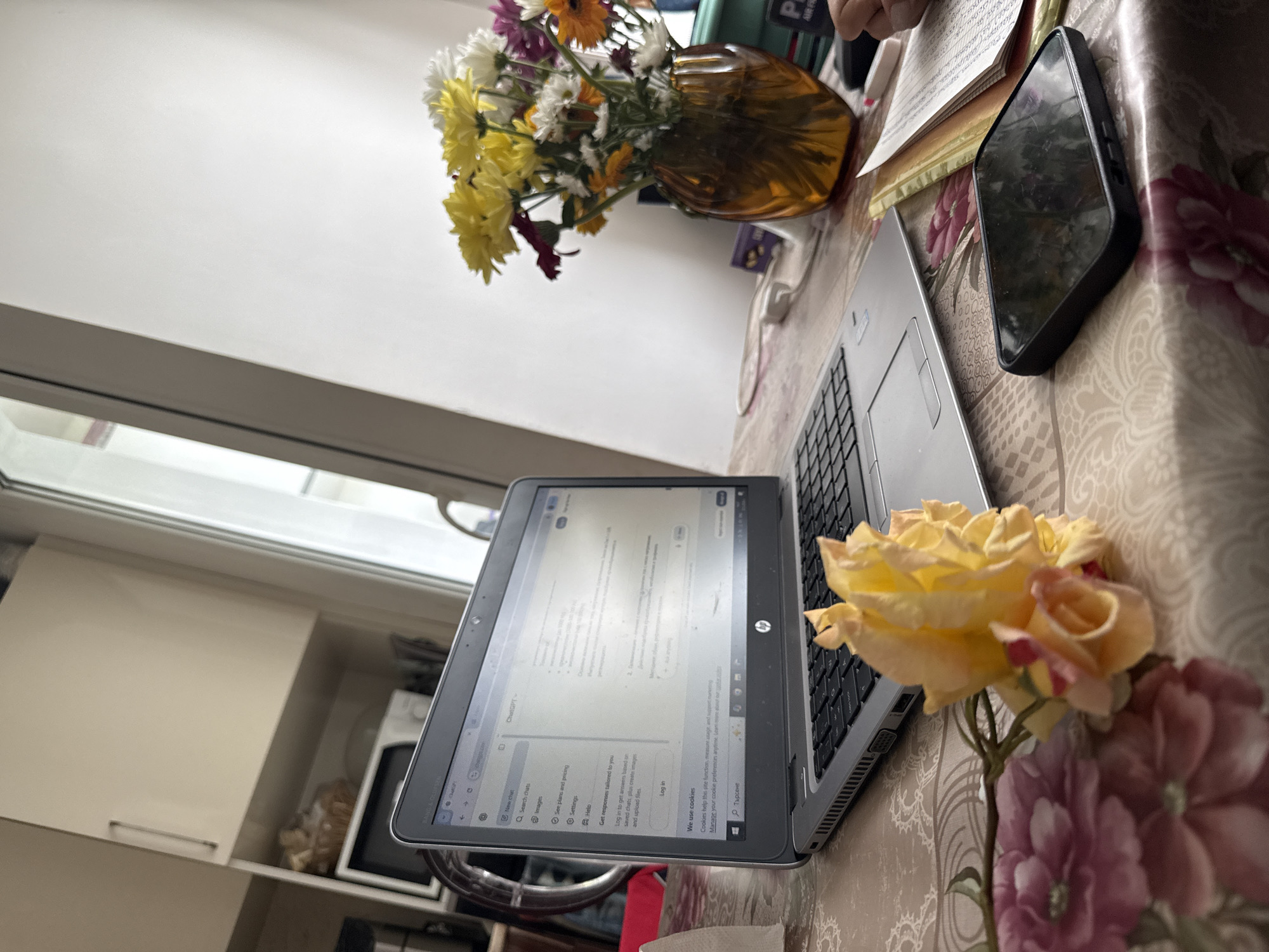 The image shows a laptop on a table with a floral tablecloth. Next to the laptop is a bouquet of flowers in a vase and a phone. The laptop screen shows the ChatGPT interface. A rose is also present next to the laptop. The scene is near a window and cabinets.
Since there is no clear indication of a specific location in the city of Stara Zagora, Bulgaria, I am unable to provide that information.
