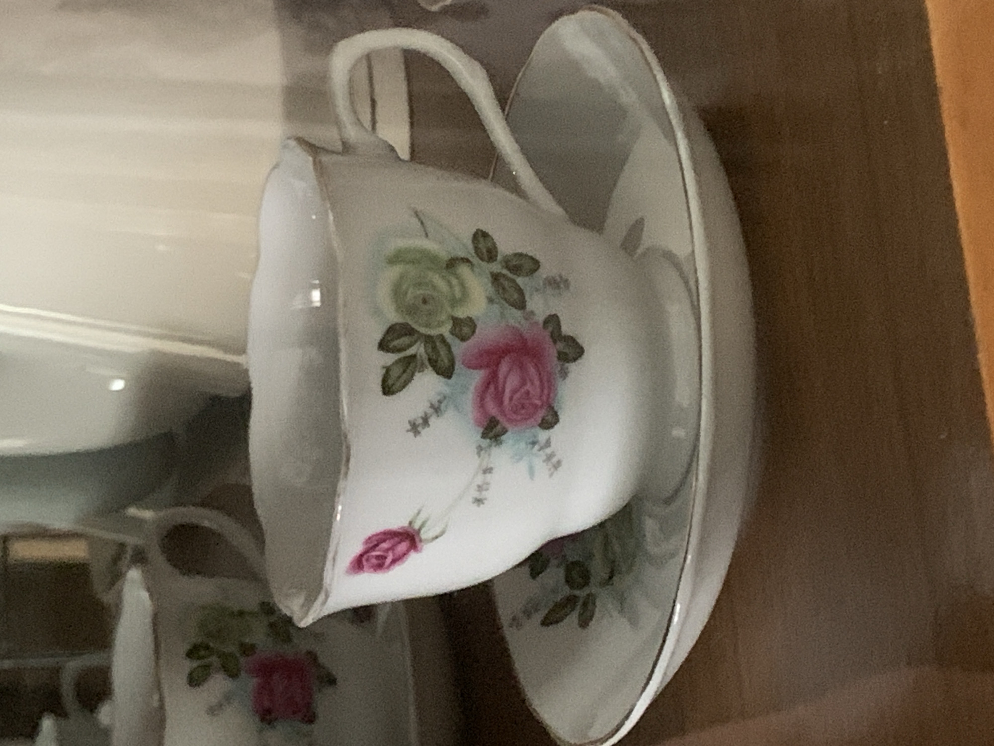 The image shows a close-up of a delicate porcelain teacup and saucer with a floral design. The teacup features painted roses in shades of pink and a lighter green, accented with small blue flowers and green leaves. The rim of the teacup and the edge of the saucer are trimmed with a thin gold band. The teacup has a gracefully curved handle. The objects are placed on a polished wooden surface. In the background, out of focus, are other ceramic items, including what appears to be a sugar bowl or creamer and part of a larger pitcher or teapot. The lighting suggests an indoor setting, possibly a display cabinet or a table. There are no people or animals visible in the image.