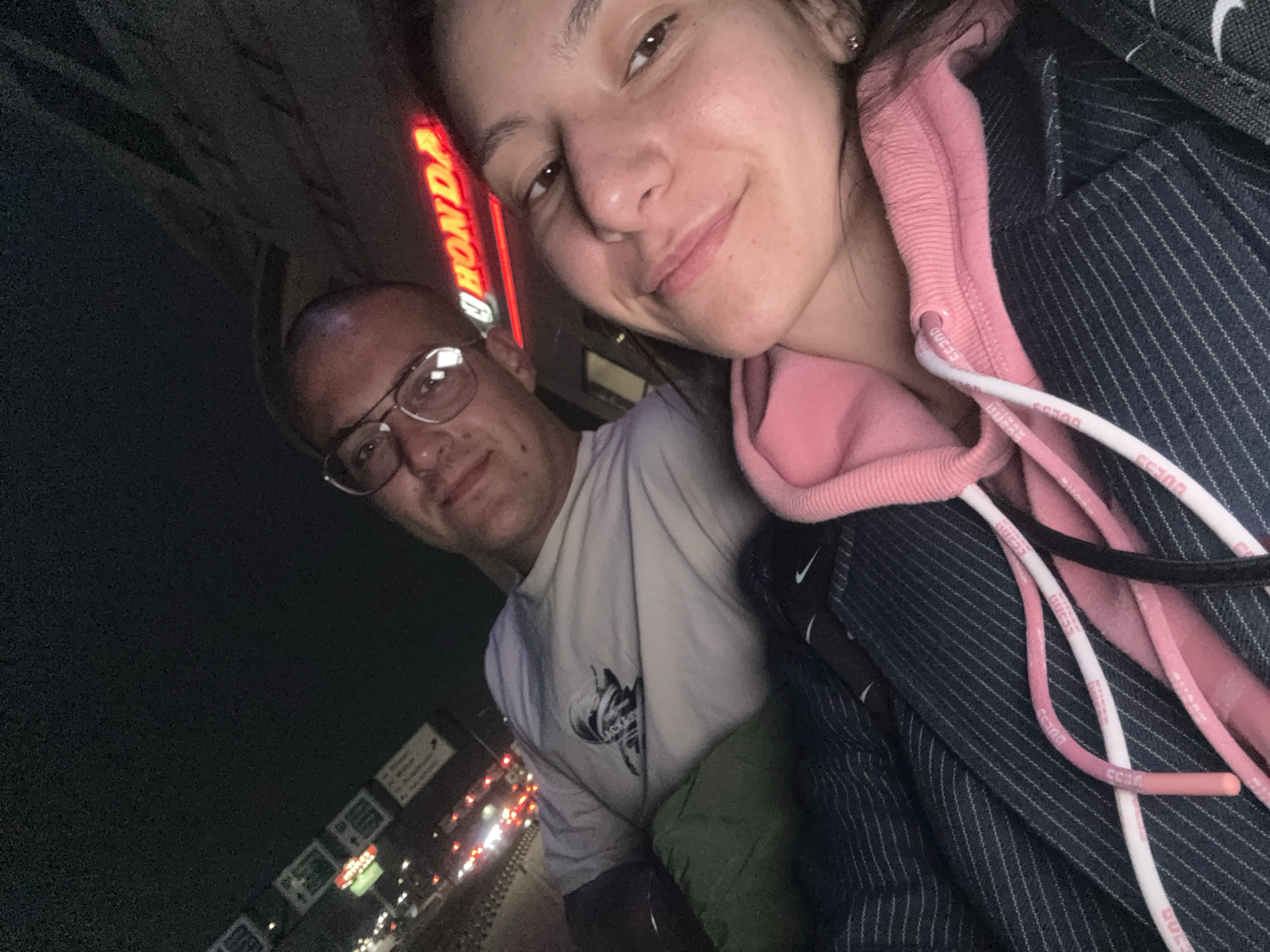 A selfie captures a woman and a man in an urban setting. The woman is in the foreground and to the right, smiling slightly at the camera. She is wearing a pink hoodie with white drawstrings marked "GUESS" and a pinstripe blazer with a black Nike swoosh logo visible. To the left and slightly behind her, a man wearing glasses and a white t-shirt looks toward the camera.
Behind the two individuals, a neon sign displaying "HONDA" in red letters is visible. Streetlights and signs are partially visible.
Based on the "HONDA" signage, the location is determined to be Honda Varna at bul. "Vladislav Varnenchik" 263, 9009 Varna, Bulgaria. The photo appears to have been taken at night or in low light.