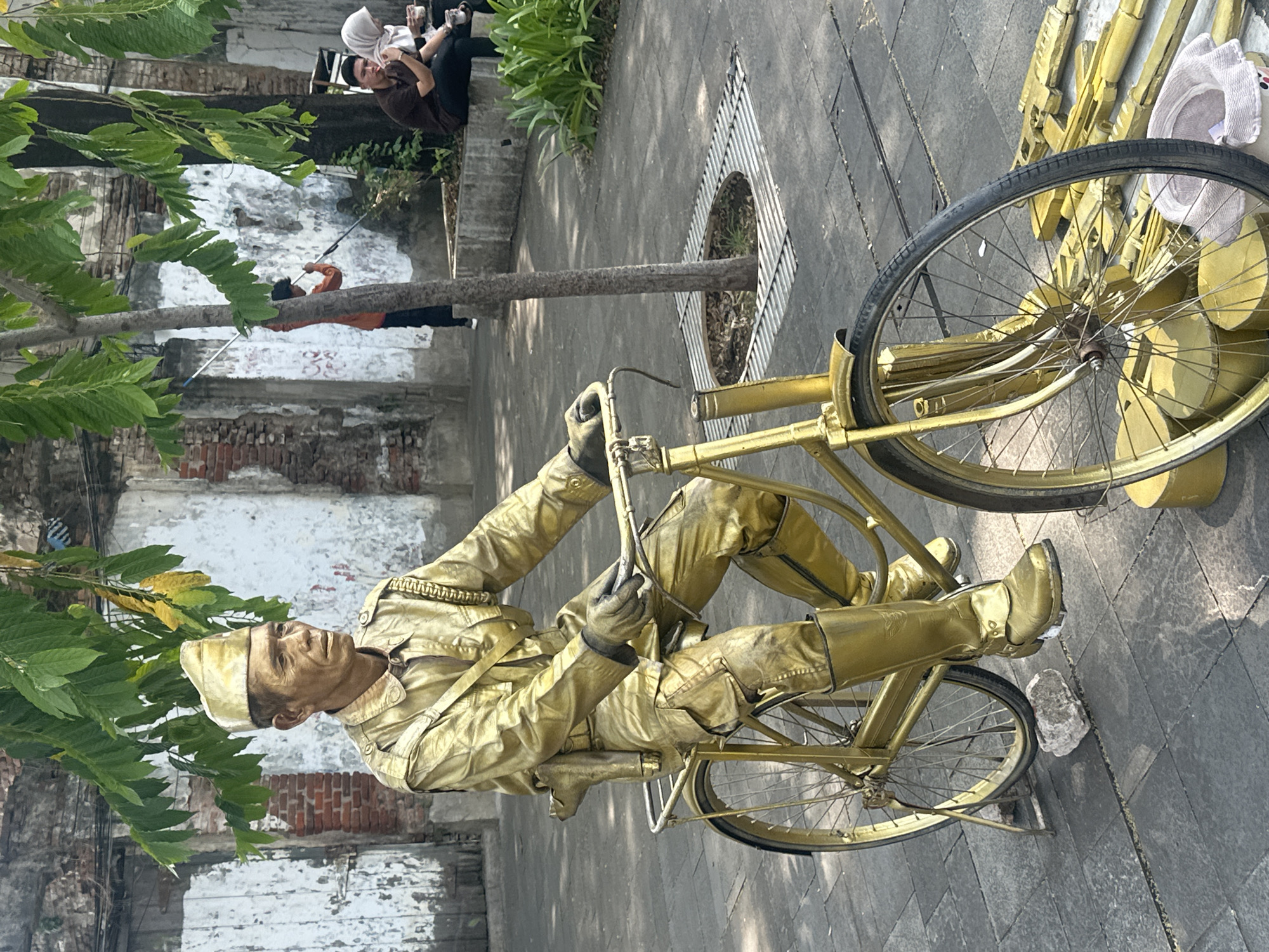 A performance artist painted entirely gold, including his clothing, is sitting still on a vintage bicycle that is also painted gold. The artist is located outdoors on a paved area with some trees and foliage in the foreground. A tree trunk runs horizontally across the middle of the image, extending from the left edge to a metal grate in the pavement. In the background, there are people and some dilapidated structures with exposed brick. The performance is taking place in West Jakarta, Indonesia.
