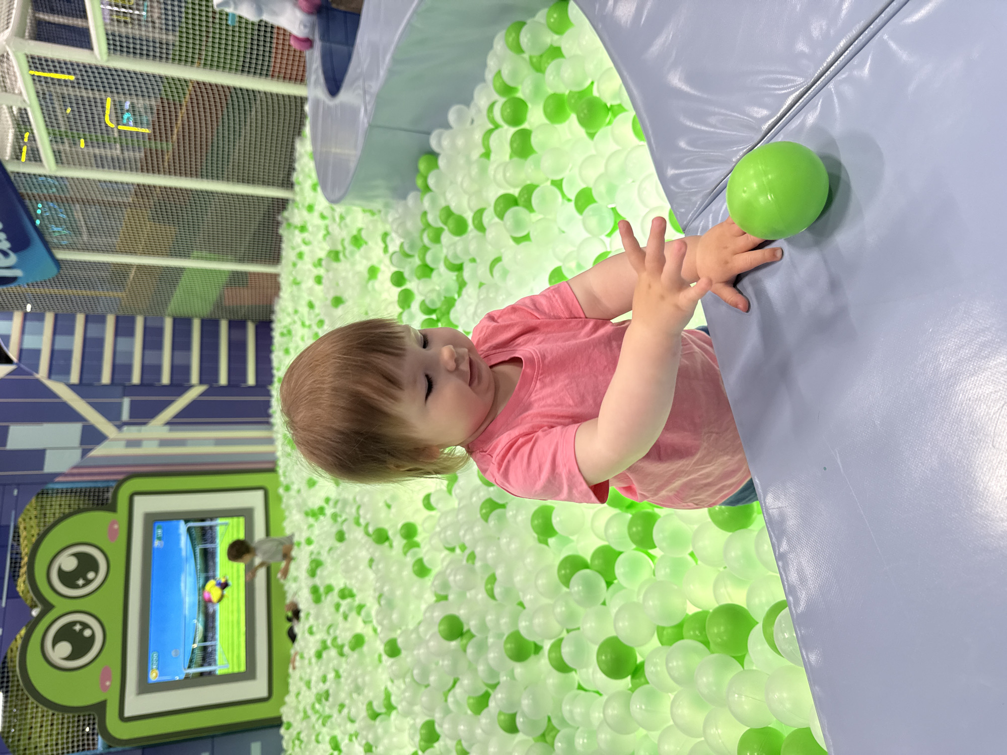 A young child in a pink shirt is in a ball pit filled with green and white balls, located inside an indoor playground. The child is reaching for a green ball resting on a blue padded surface. A screen with a cartoonish frog design shows children playing a soccer game. The playground includes netting and structures. The location of the scene is Перовское сельское поселение, Ukraine.