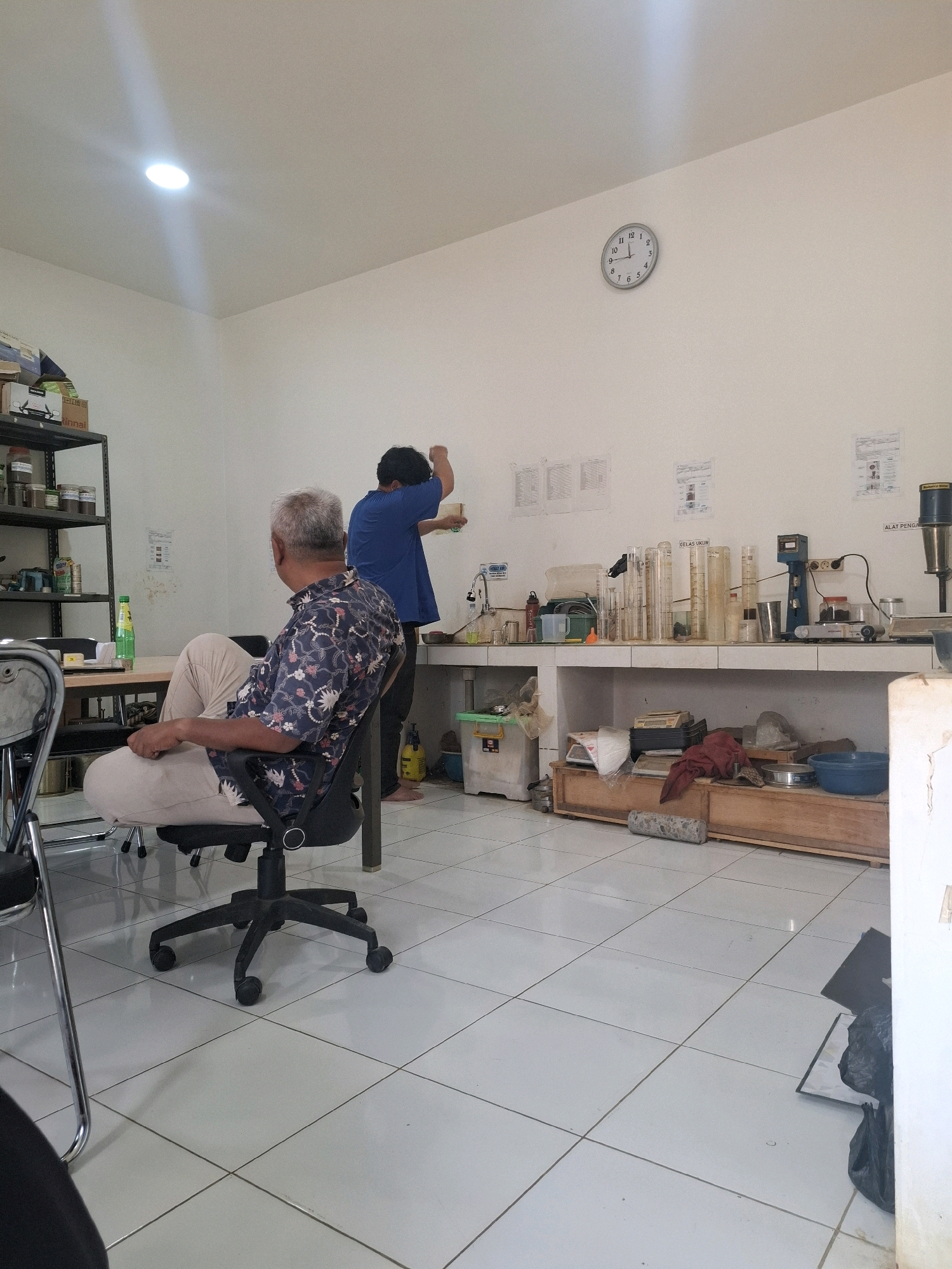 The image depicts an indoor scene, likely a laboratory or workshop, located in Ciheulang Tonggoh, Indonesia. In the foreground, a man with graying hair is seated in a black office chair, facing away from the camera and towards a table on his left. He is wearing a patterned short-sleeved shirt and light-colored pants.
In the background, another person wearing a blue t-shirt is standing and appears to be interacting with equipment on a counter. The room is illuminated by ceiling lights, and a white wall features a circular clock. The counter is equipped with various scientific apparatus, including graduated cylinders, beakers, and what appears to be a centrifuge or similar device. Below the counter, a wooden cabinet displays several items, including scales, containers, and fabric. The floor is tiled with white squares. Shelving units are visible on the left side of the room, holding jars and bottles. Other objects in the environment include a green bottle, a yellow spray bottle, and trash bags. The overall impression is one of a functional workspace with ongoing activity.