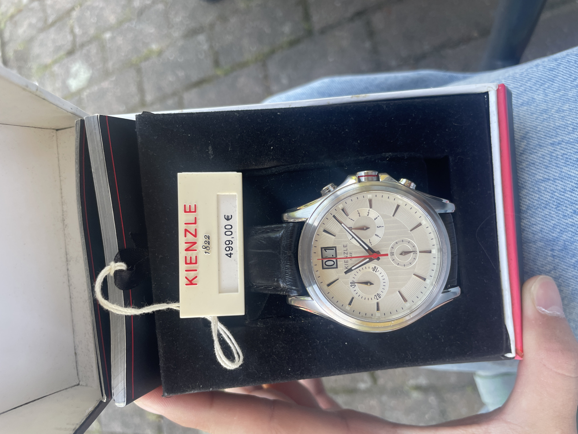 The image shows a boxed Kienzle 1822 wristwatch, possibly for sale, with a price tag of 499.00 EUR. The watch has a silver-tone casing, black leather strap, and a light-colored face. The date on the watch is "01". The background includes a person wearing light-colored denim, and an out-of-focus pavement or pathway. This image could be from Wienhausen, Germany.