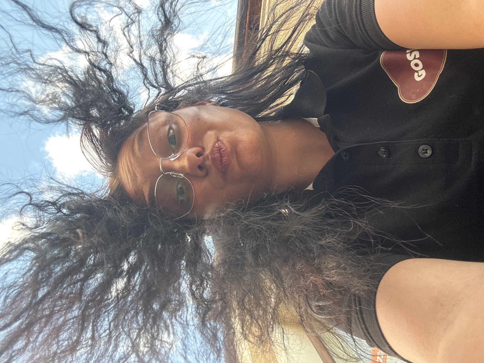 A woman with dark hair, wearing glasses and a black Costa Coffee branded shirt, is positioned looking upwards towards the sky. Her hair is disheveled, appearing to be caught in the wind. The background includes a partly cloudy sky and a glimpse of a building structure. The image suggests an outdoor location, possibly in an urban area, but the specific location in Sofia, Bulgaria is not evident.