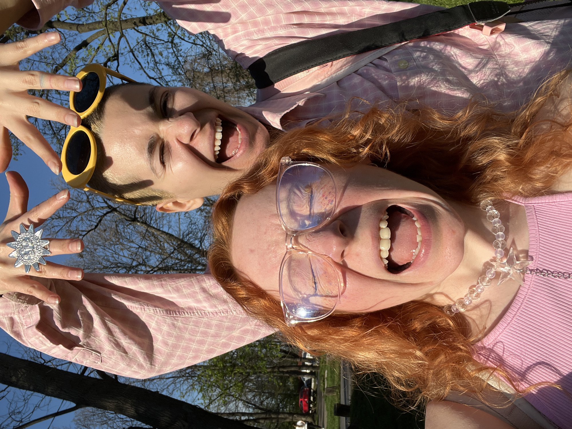 Two individuals are the subjects of the photo. The individual on the left has short blonde hair and is wearing yellow sunglasses. They are making hand gestures toward the camera, displaying a large silver ring on their finger. The individual on the right has red hair and is wearing clear-framed glasses and a beaded necklace with a star pendant. Both individuals are wearing pink tops and have their mouths open in expressions of laughter or excitement. The background suggests an outdoor setting with trees and a blue sky. There are hints of urban infrastructure like roads, cars, and perhaps park furniture. The location within Belgrade, Serbia is indiscernible without further visual markers or context.