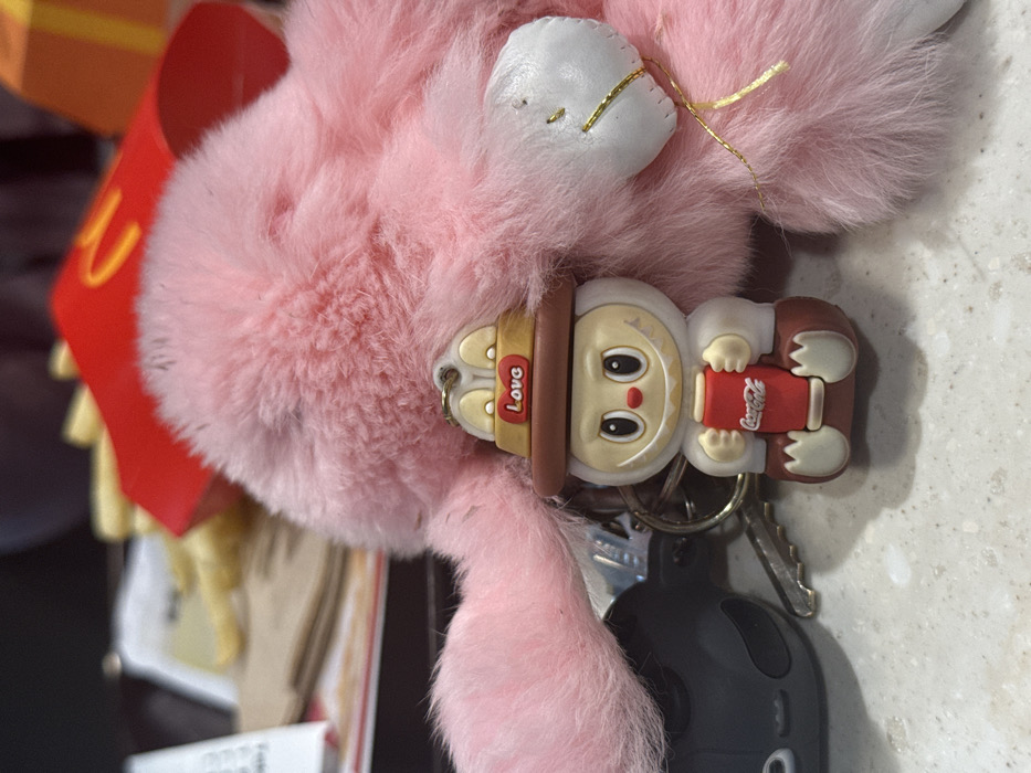 A fluffy pink bunny keychain with white ears and a smaller bunny figurine holding a Coca-Cola can are on a keyring. A car key is also attached to the keyring. In the background are McDonald's fries and other items. The surface underneath is light-colored.