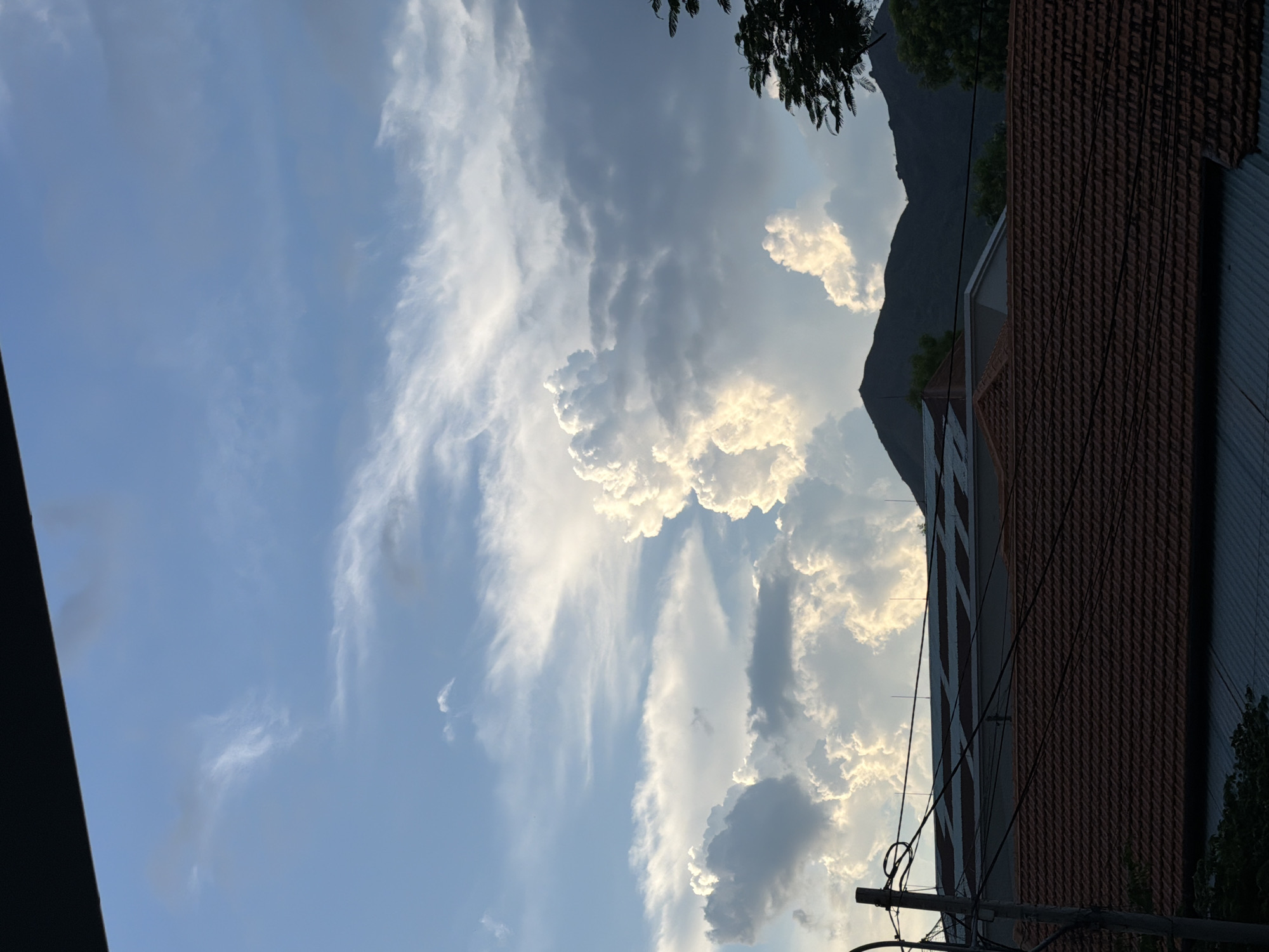 A low-angle, wide shot captures a view of the sky and rooftops in Bac Nha Trang Ward, Vietnam. The sky is filled with a dramatic mix of clouds, with bright, voluminous cumulus clouds illuminated by sunlight, contrasting with darker, grayer cloud formations. Wispy cirrus clouds streak across the upper portion of the sky. Below the clouds, the silhouettes of dark, jagged mountains are visible in the distance. In the foreground, a utility pole with tangled wires stands to the left. Several rooftops with red tiles and some with corrugated metal sheeting are visible. Trees and foliage are present on the right side of the frame and at the bottom. The lighting suggests it is either sunrise or sunset, with the sun's rays backlighting the clouds. No people or specific actions are depicted.
