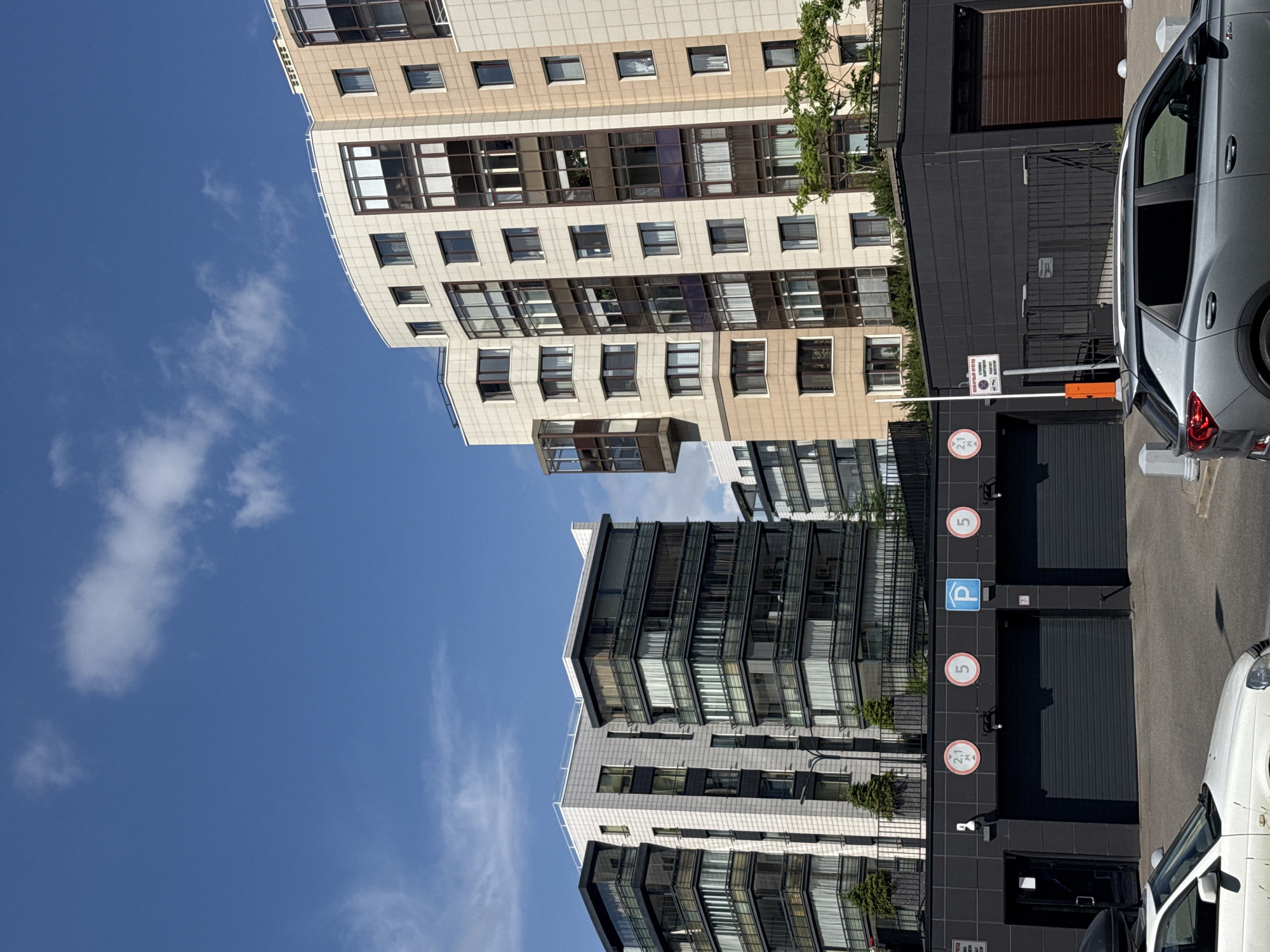 This is an outdoor, eye-level shot of a modern residential building complex with a clear blue sky overhead. The buildings are multi-story structures with a mix of beige and dark gray facades, featuring numerous windows, some with balconies and glass enclosures. The architecture appears contemporary.
In the foreground and right side of the image, a gray car is parked, and partially visible is a white car. Behind the cars, there's a dark-colored entrance to a garage with rolling doors. Several signs are visible on the dark wall next to the garage doors, including parking signs with a blue "P" symbol and circular signs with the number "5" in white on a red background, suggesting parking levels or restrictions. Another circular sign with "2.1 M" is also visible, likely indicating a height limit. A security barrier arm is positioned across what appears to be an entrance or exit.
The sun appears to be high in the sky, casting shadows, suggesting it is daytime, likely late morning or afternoon. The weather appears to be clear and sunny with a few wispy clouds scattered in the sky. No people are clearly visible in the frame, focusing the attention on the architecture and the vehicles. The scene has a clean, organized, and urban feel, possibly a residential complex or a modern development. There is some greenery, including small trees, visible near the buildings and along the dark facade.