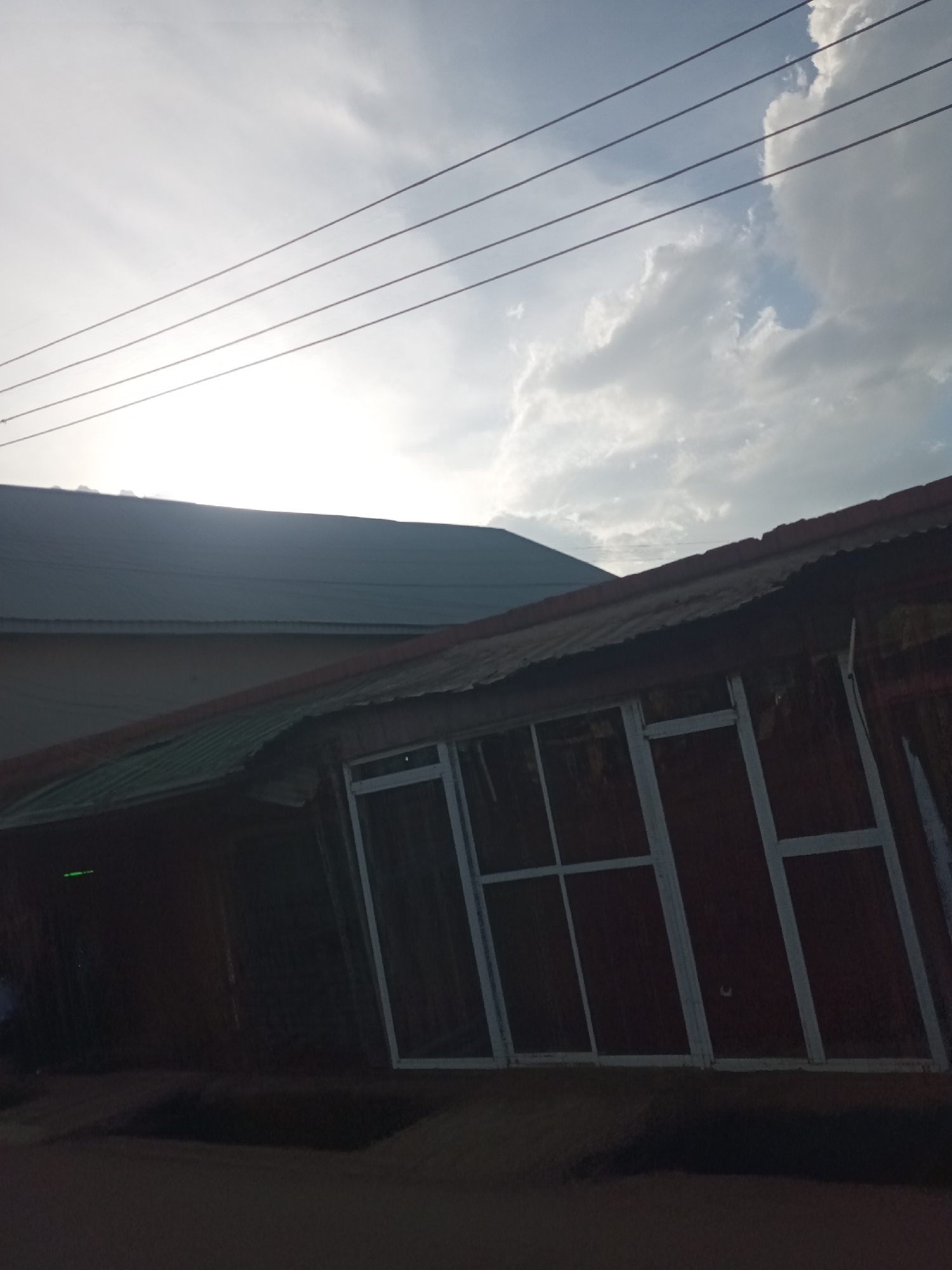 The photograph captures a daytime outdoor scene in an urban or semi-urban setting, consistent with Jos, Nigeria. The image has a notable tilt, framing buildings against a bright, cloudy sky intersected by utility wires.
In the foreground, a building with a dark, reddish-brown exterior and a corrugated metal roof is prominent. The lower section of this structure is heavily shadowed, almost appearing as a silhouette, though its features include a set of light-colored, possibly white, window or door frames divided into multiple rectangular sections. A faint green light or reflection is visible on the far left of this building's darkened facade.
Behind this foreground building, another structure with a darker grey, possibly corrugated metal roof can be seen. This building appears larger, with its roofline extending horizontally across the frame.
The upper portion of the image features a bright, cloudy sky. An intense light source, likely the sun, is positioned in the upper-left, causing a significant overexposure in that area. White and grey clouds are scattered across the sky, with hints of blue peaking through, particularly towards the upper right. Four distinct, thin, parallel lines, consistent with power or telephone wires, stretch horizontally across the sky.
The strong contrast between the bright sky and the heavily shadowed buildings suggests either a high sun angle or an exposure setting prioritizing the sky. No people are visible, and no specific activities or events are depicted. There is no visible text in the image. The scene presents a stark yet common view of local architecture and infrastructure under bright daylight conditions.