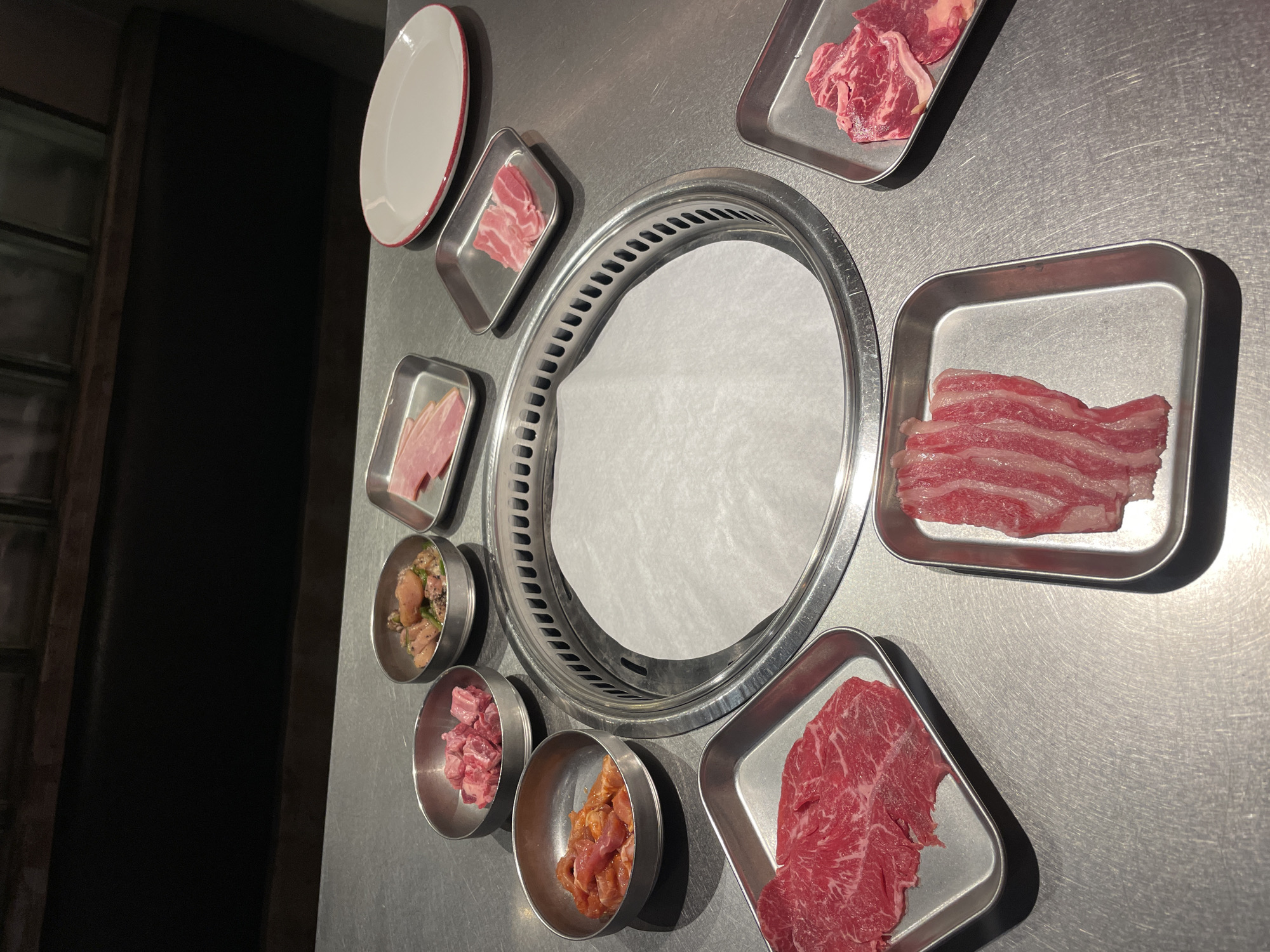 The image depicts a tabletop at a Korean BBQ restaurant, likely in Barcelona, Spain. A circular grill sits in the center, lined with white paper. Surrounding the grill are various metal trays and bowls containing different types of uncooked meat, including thinly sliced beef, cubed meat, and marinated chicken. A white plate with a red rim is also present. The surface beneath is a stainless-steel counter. The scene appears to be a setup before cooking.