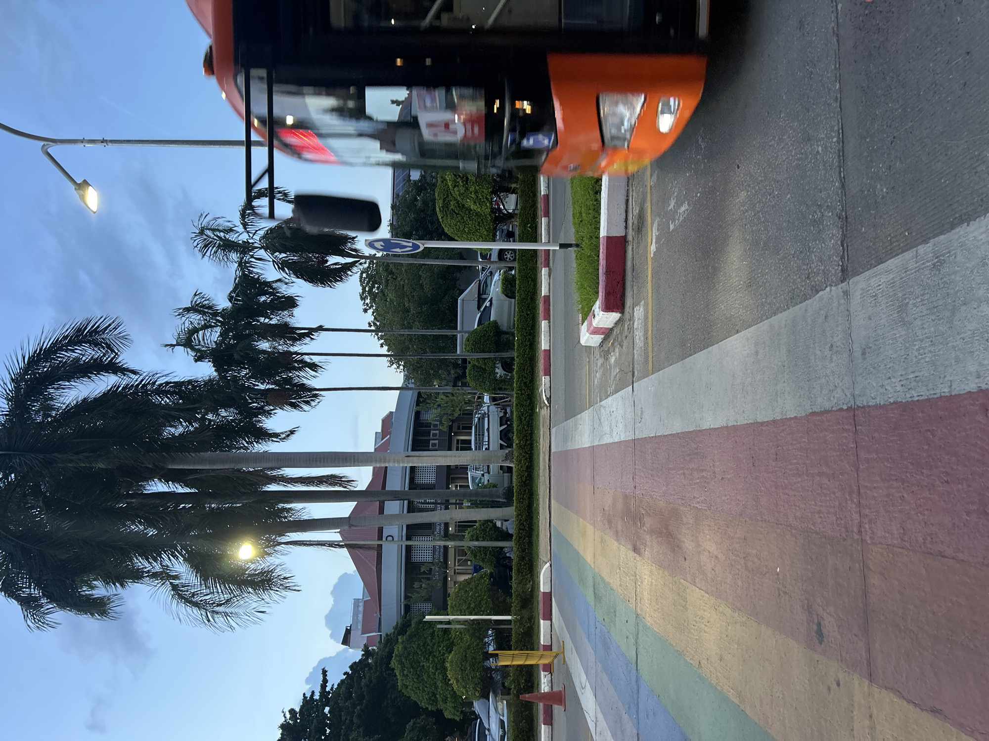 The image depicts a street scene in Tha Khlong Town Municipality, Thailand, at dusk. The foreground features a rainbow-colored crosswalk, with stripes of purple, blue, green, yellow, and pink. To the right, an orange bus is in motion, its front visible, with its headlights on. The bus has a black interior and an orange-tinted windshield. A blurry black object hangs from the bus's windshield. A streetlamp with an illuminated bulb stands behind the bus. Palm trees with lush green foliage are scattered throughout the scene, some reaching towards the twilight sky. In the background, a building with a red roof and light-colored walls, featuring several windows and vertical columns, is visible. Several parked cars and a white pickup truck are also present. The environment suggests an urban setting with vegetation.