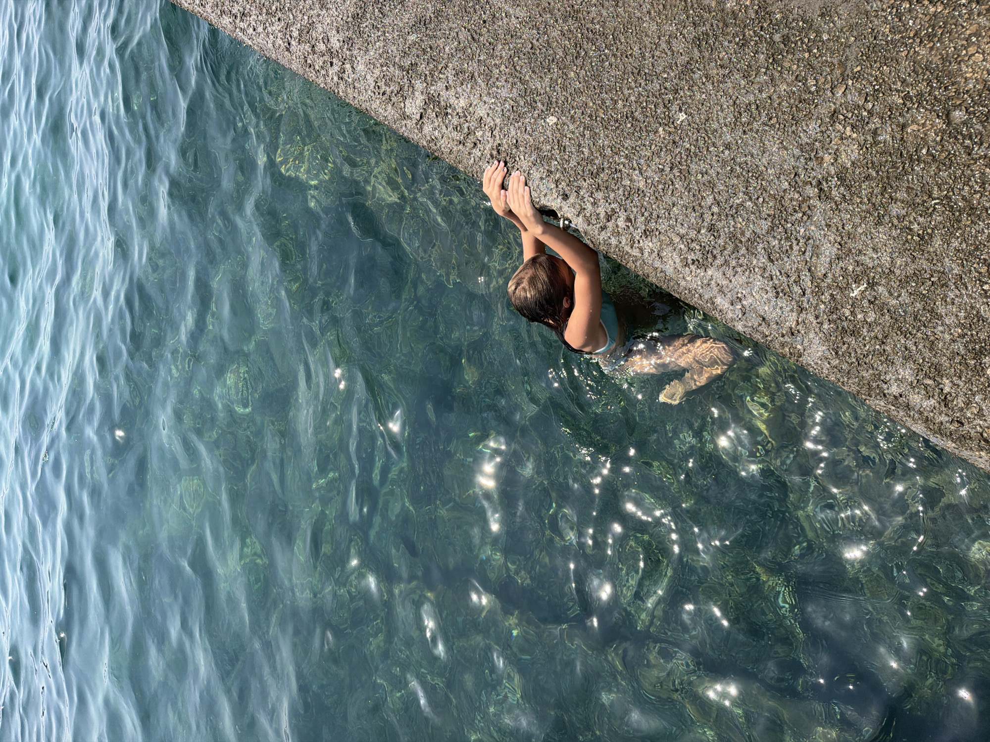 The image shows a person in clear, turquoise water next to a textured concrete structure. The person, a young individual with blonde hair, is submerged in the water up to their chest and is holding onto the edge of the concrete with their hands. Sunlight glints off the water's surface, creating shimmering reflections. The scene appears to be outdoors on a sunny day, likely a warm climate given the activity. The concrete edge suggests a dock, pier, or wall. The water is translucent, allowing a view of the submerged bottom in some areas. The overall impression is one of a refreshing dip in a natural body of water. The location context suggests Budva, Montenegro.