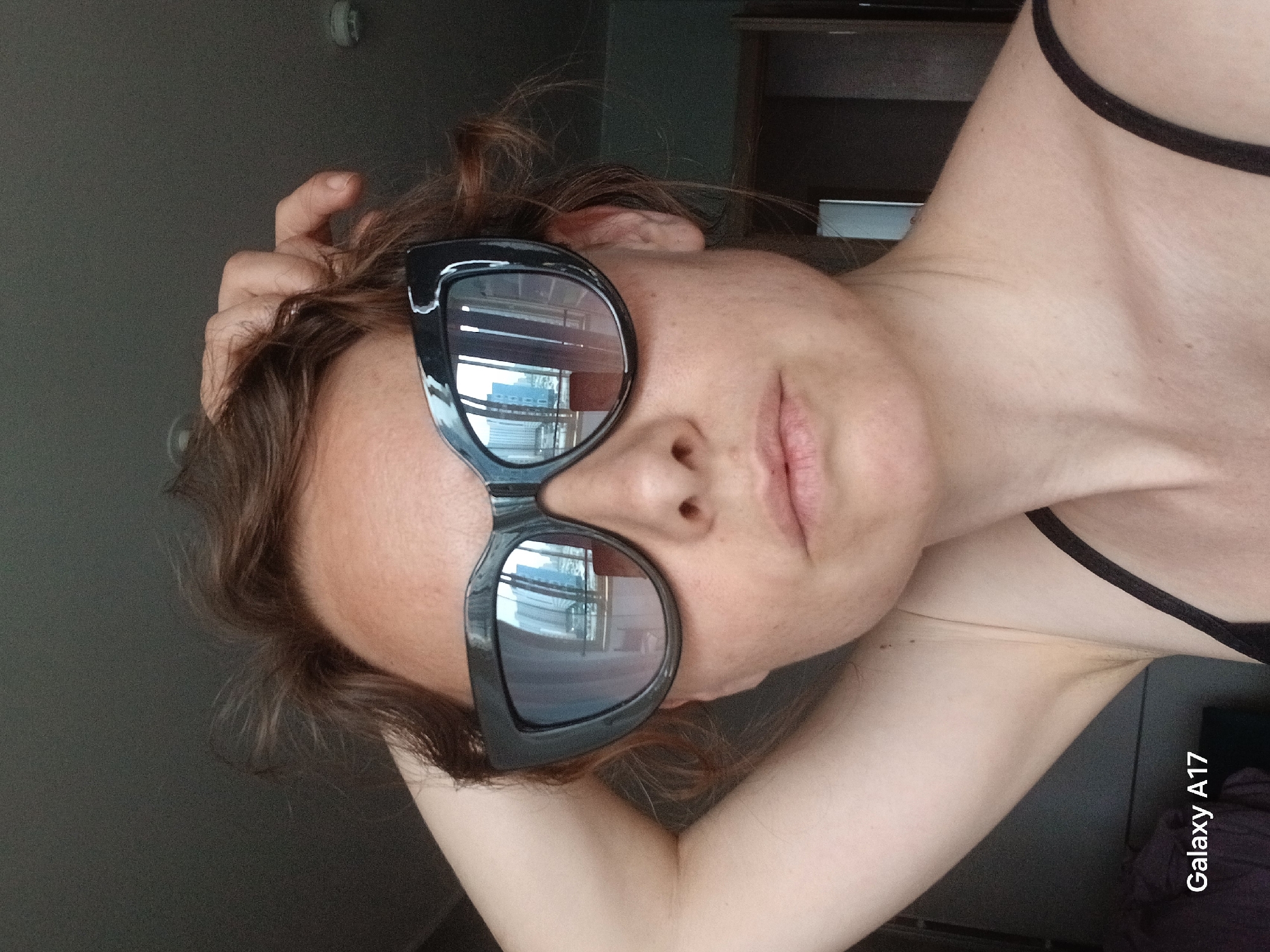 A woman with brown curly hair is wearing black sunglasses. She has her hand on her head. The reflection in her sunglasses shows buildings in Yekaterinburg, Russia. The lighting is soft. The image is rotated. A black tank top strap is visible. A "Galaxy A17" watermark is in the bottom right corner.