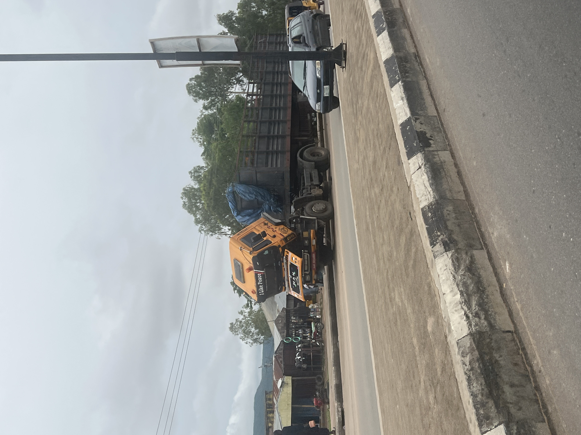 The image depicts a road scene in Jalingo, Nigeria, characterized by a tilted yellow truck with "I Like Peace" and "MAN" written on its cabin. The truck's cabin is significantly angled upwards, suggesting an accident or improper loading. The truck's trailer is filled with greenery, covered partially with a blue tarp. Beside the truck, a dark sedan is visible on the road, and a yellow three-wheeled vehicle is in the distance. The background features a row of shops with various items displayed, and beyond that, a line of trees and hills under an overcast sky. A lamppost stands on the right side of the image, and a person in dark clothing is walking on the sidewalk to the left. The road has a concrete barrier with black and white markings.