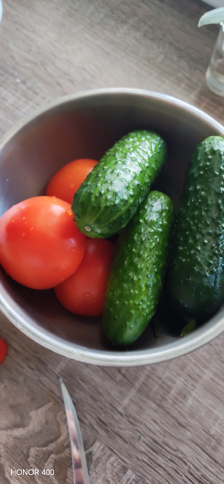 A top-down view reveals a silver metal bowl containing three cucumbers and three tomatoes. A knife blade is partially visible at the bottom left of the frame. The bowl is sitting on a wooden surface. A portion of a glass vase is visible at the upper right of the frame. The bottom of the image states "HONOR 400." The location of Noviy, Russia, is not discernible from the image.