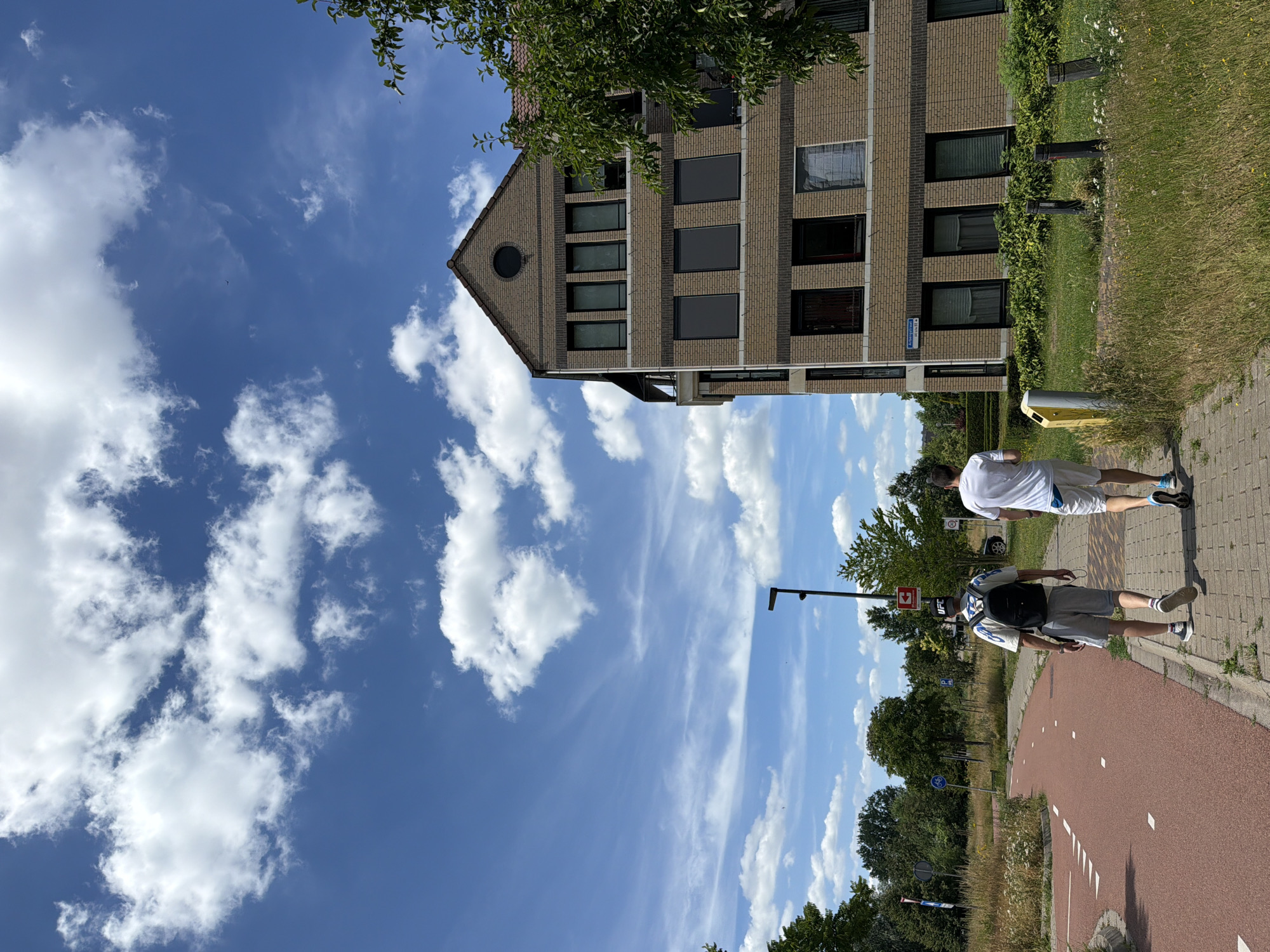 Two males walk away from the camera on a brick sidewalk next to a red bike path. The male in front is wearing a backpack, grey shorts, and a white t-shirt with blue and grey patterns. He is also wearing a black cap with "UFC" written on it. The male behind him is wearing white shorts and a white t-shirt, with blue athletic shoes. They are walking on Alberingstraat in the city of Helmond, Netherlands. To the right of the sidewalk is a multi-story brick building with dark-framed windows. To the left of the bike path are trees and vegetation, and a blue street sign with white lettering. The sky is blue with white, wispy clouds.