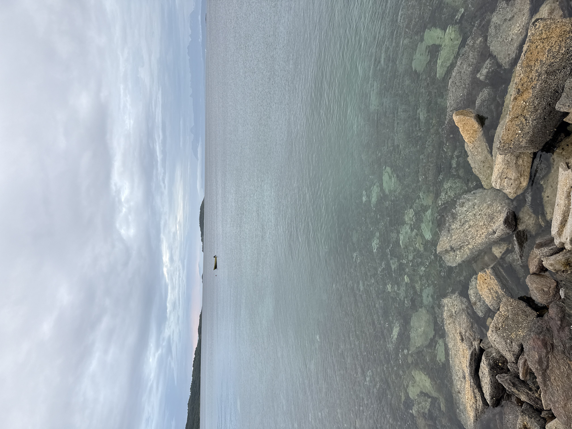 A wide, high-angle shot captures a serene, overcast seascape. In the foreground, a collection of large, weathered rocks sits at the edge of the water, some partially submerged. The water is clear and shallow near the shore, revealing submerged rocks and pebbles, before transitioning to a deeper, greyish-blue expanse. In the middle distance, a small, yellow boat floats serenely on the calm water. The horizon is marked by a low-lying, tree-covered island to the left, and in the far distance, a range of misty mountains can be seen against the backdrop of a cloudy sky. The overall impression is one of tranquility and natural beauty. This scene is consistent with the coastal landscape found in and around Harstad, Norway.