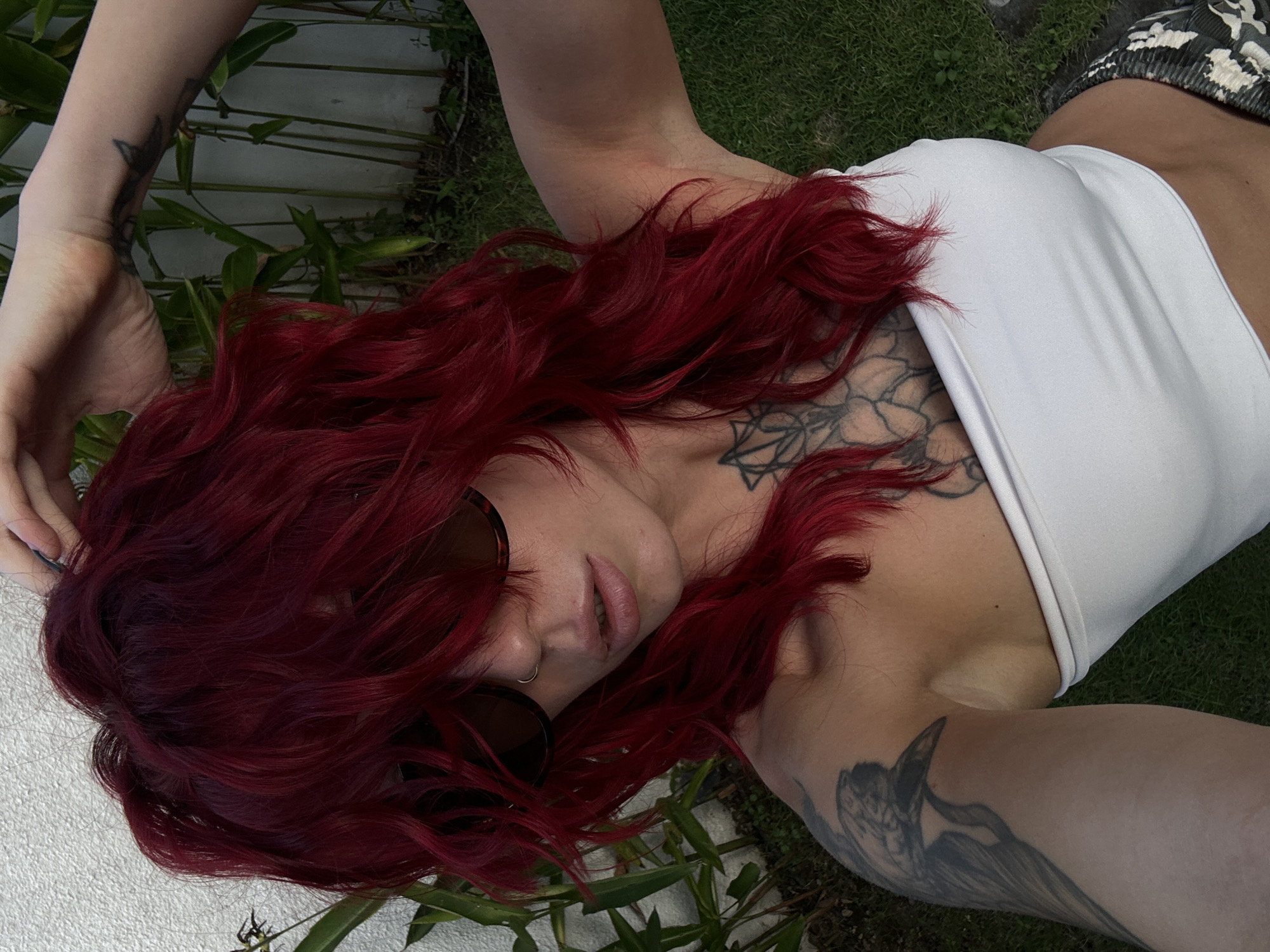 A medium shot captured from a high angle shows a woman with bright red, choppy, shoulder-length hair, wearing sunglasses and a white strapless top. She is touching the top of her head with her right hand, and her left arm is extended to take the photo. She has a nose ring and several tattoos visible on her arms and chest. The background consists of lush green grass, some foliage, and a white wall. The lighting suggests an outdoor setting, possibly during the day. The woman's expression is partially obscured by her sunglasses and hair, but her mouth is slightly open. The overall impression is of a casual, candid moment. The scene appears to be an outdoor space, possibly a garden or patio, in a tropical or sub-tropical climate given the vegetation.