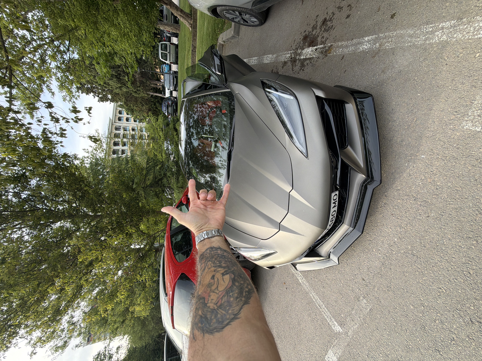 A person's tattooed arm, making a rock 'n' roll hand gesture, is in the foreground of the image, obscuring the front of a metallic grey sports car. The car has its driver's side door open and is parked in a lot with white parking lines. In the background, a line of other vehicles, including a white van and several dark-colored cars, are visible, suggesting a parking lot or roadside. Lush green trees with dense foliage line the left side of the image, and a modern building with multiple windows and balconies is partially visible behind the trees. The pavement is asphalt, with some damp patches indicating recent rain or washing. The time of day appears to be afternoon, with the lighting casting subtle shadows. The sky is overcast, suggesting cloudy weather. The license plate of the sports car is partially visible and reads "BG 480 MO". The overall scene depicts a casual outdoor setting, likely a parking area in Varna, Bulgaria.
