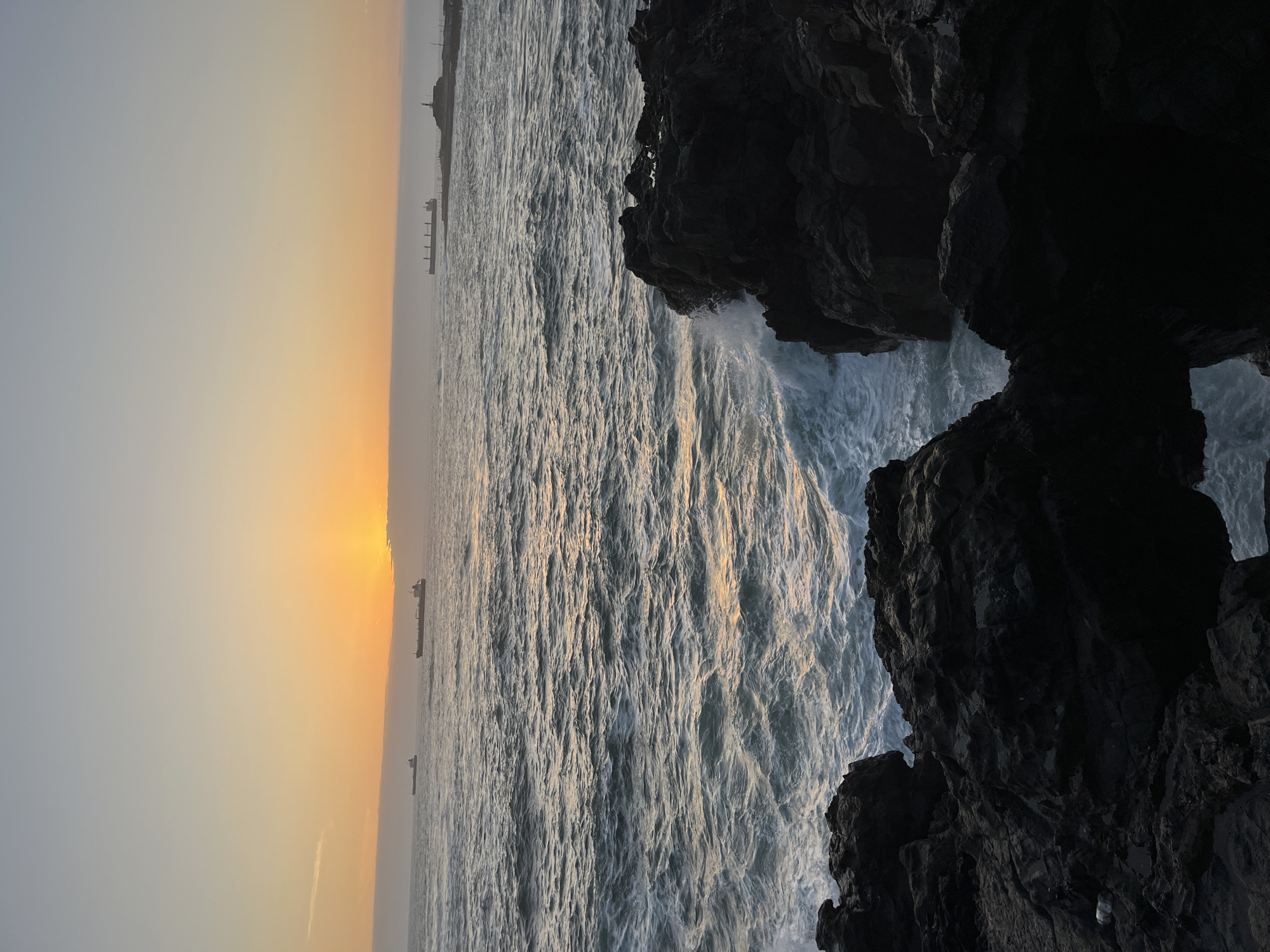 The image depicts a seascape in Arica, Chile, at sunset. Dark, jagged rocks are in the right foreground with waves crashing against them. The sun is setting over the ocean in the background, casting a golden glow on the water and sky. There are several ships visible on the horizon.