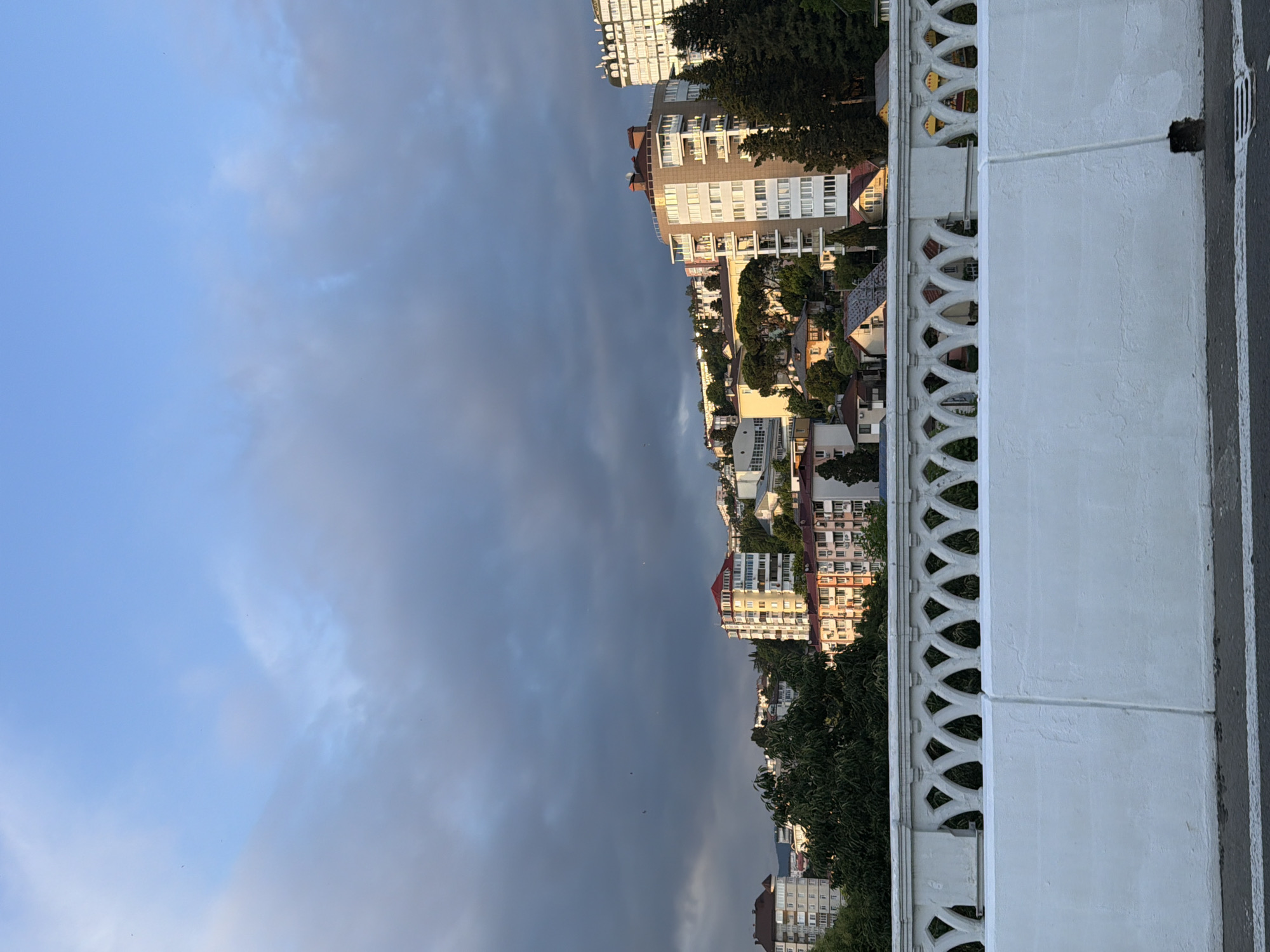 The image depicts a cityscape of Sochi, Russia, with a focus on buildings and a cloudy sky. The perspective is from an elevated vantage point, looking down over a series of apartment buildings nestled amongst lush green trees. The buildings vary in height and architectural style, with many featuring balconies and light-colored facades. Some structures appear modern, while others have a more traditional design with red-tiled roofs.
In the foreground, a white decorative railing with an intricate, repeating pattern runs horizontally across the frame, separating the viewer from the city below. To the right of the railing, a paved surface, possibly a sidewalk or road, is visible with white lines marking lanes or boundaries.
The sky is a prominent feature, dominated by heavy, dark clouds that obscure much of the sun, suggesting overcast or stormy weather. Patches of lighter sky are visible, hinting at a break in the clouds or the approach of a storm. A few small, dark shapes, possibly birds, are faintly discernible in the sky. The lighting suggests it is either early morning or late afternoon, with the sun's rays attempting to break through the clouds, casting a soft, diffused light on the scene. No people are visible in the image, and no text is present.
