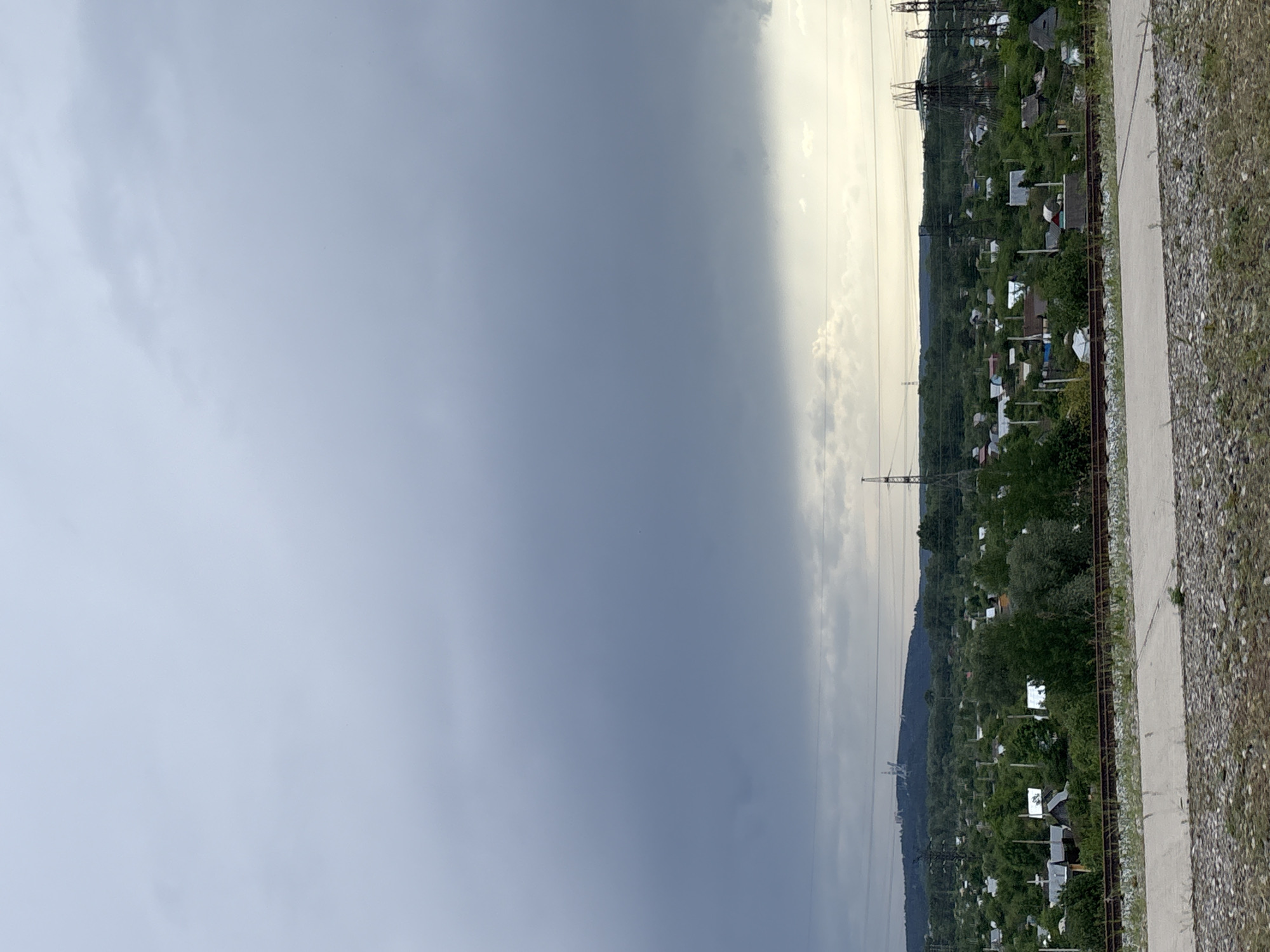 The image depicts a view in the city of Chaikovsky, Russia. A heavy, dark storm cloud dominates the left side of the frame, contrasting sharply with the brighter, slightly hazy sky on the right.
On the right side of the image, a landscape with dense vegetation and small buildings, likely residential or garden plots, is visible. Power lines run through the scene, with pylons and wires creating a visual grid against the sky and landscape. A narrow path or road runs along the edge of the frame, with a gravelly or stony shoulder. The overall impression is of a landscape on the edge of a city or town, caught between an approaching storm and a calmer, brighter area.