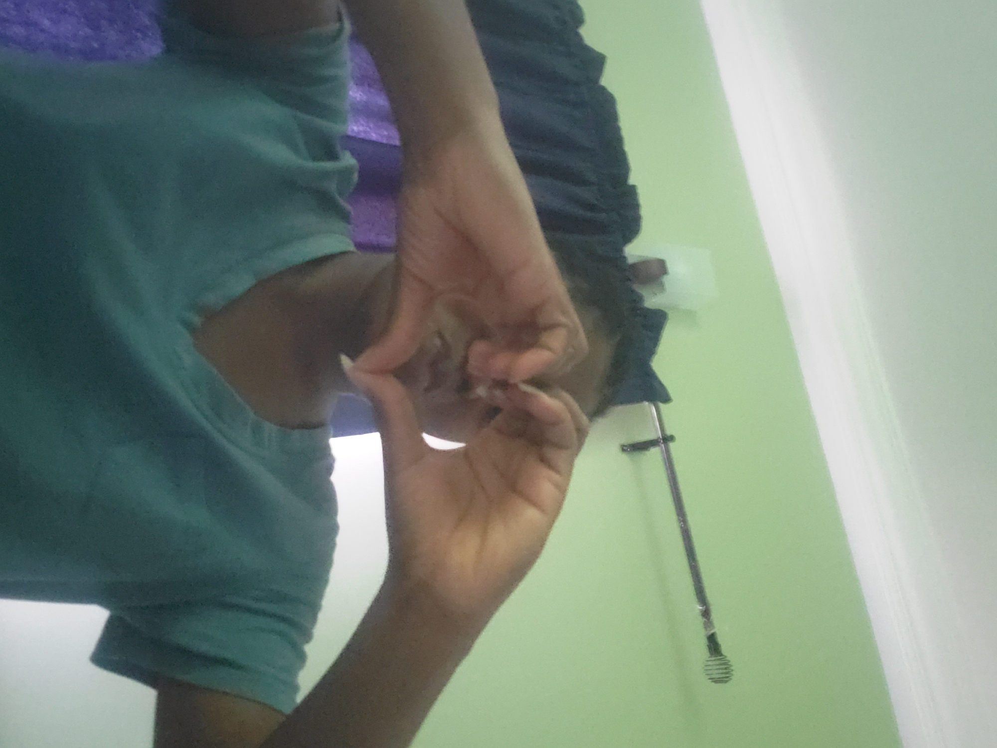 A person wearing a teal t-shirt and a dark blue ruffled garment is indoors. The person is making a circle with their thumb and forefinger, seemingly close to their face. A chrome metal shower head and mount are visible against a pale green wall. The location is likely a bathroom. The image does not provide enough information to confirm the location in Barden Yana, Nigeria.