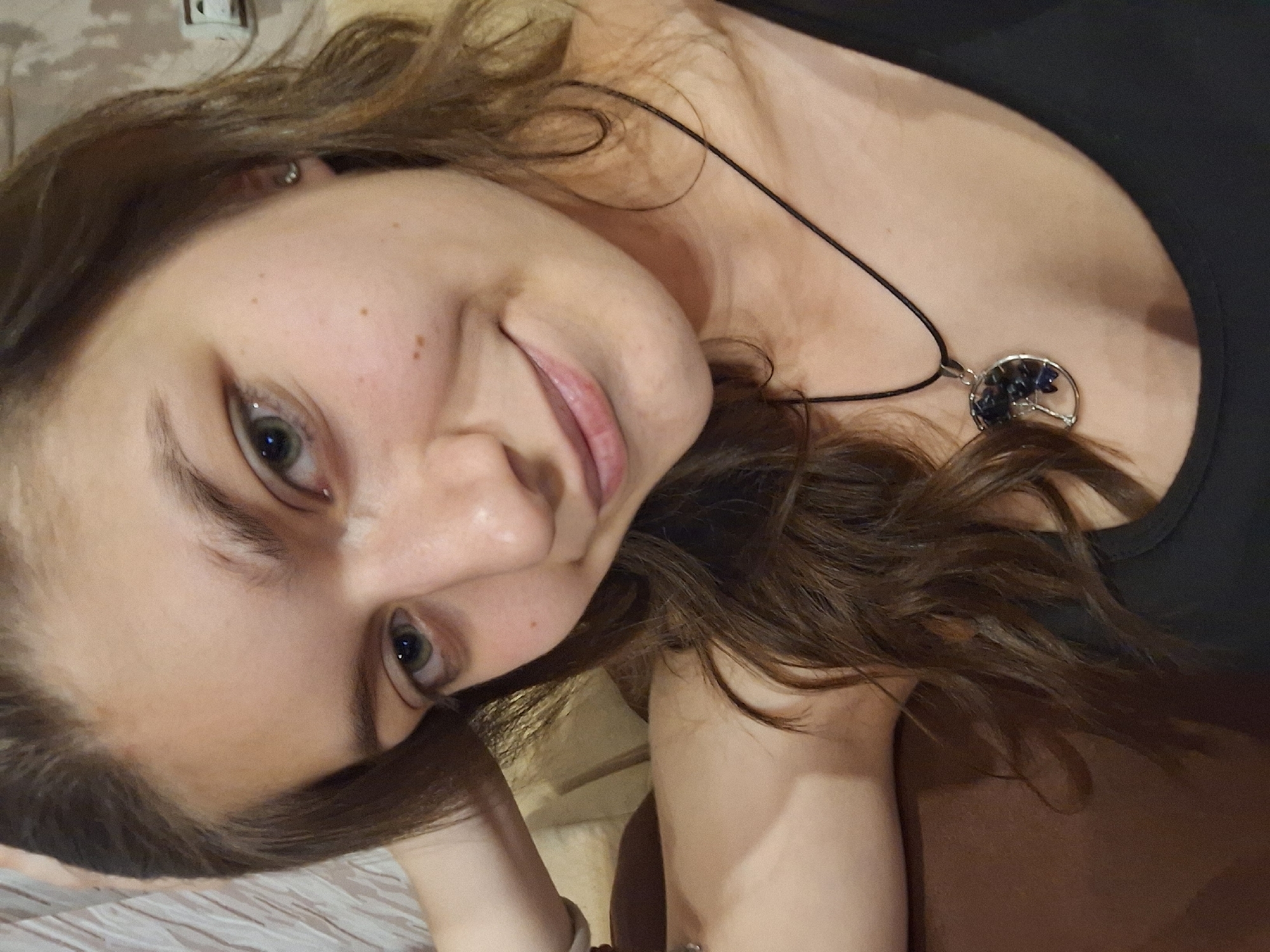 A woman with brown hair and light skin is positioned in the foreground. She is smiling and wearing a black top. A necklace with a circular pendant containing blue stones is visible around her neck. The background is out of focus. There is no evidence to indicate that the picture was taken in the city of Panagyurishte, Bulgaria.