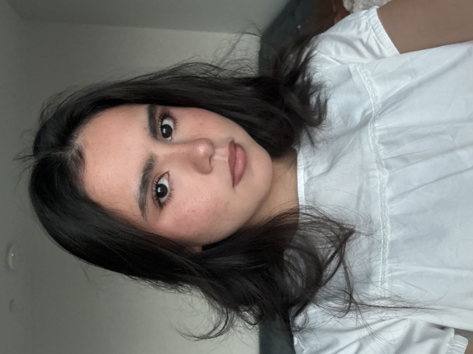A young woman with dark, wavy hair is shown from the shoulders up, facing the camera directly. She is wearing a white, short-sleeved blouse with a ruffled neckline. Her makeup includes blush on her cheeks and light-colored lipstick. The background is a plain, neutral wall. The lighting suggests an indoor setting, possibly a home. There is no indication of the location being in Austin, United States of America.