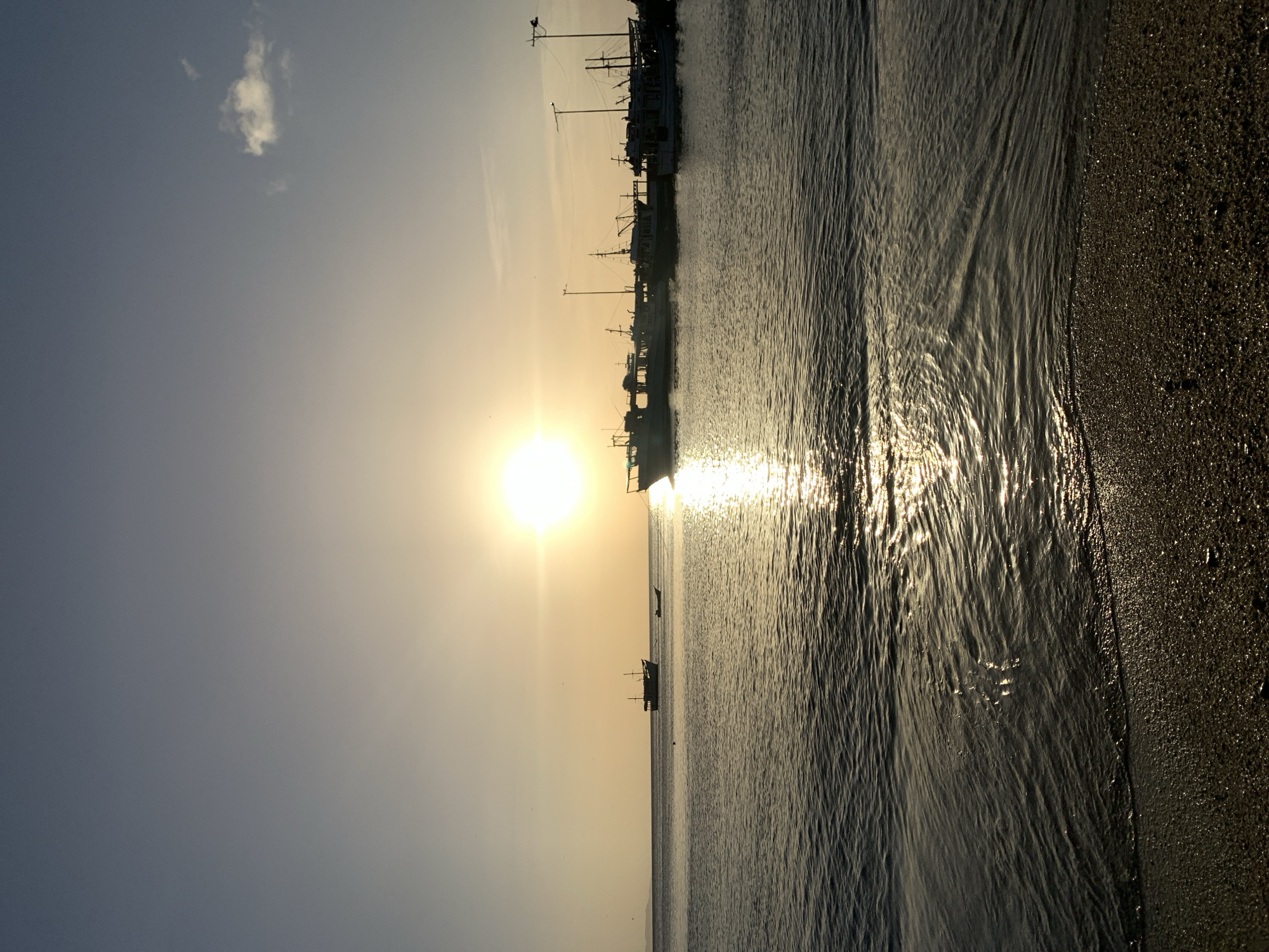 This image captures a tranquil sunset scene in Juan Griego, Venezuela. The sun, a bright orb low on the horizon, casts a golden glow across the water and sky. Several boats are docked in the harbor, their silhouettes sharp against the illuminated backdrop. The gentle waves lap at the sandy shore in the foreground, reflecting the sun's warm light. A few small clouds drift in the vast sky, adding texture to the serene atmosphere. The overall impression is one of peacefulness and natural beauty at the end of the day.