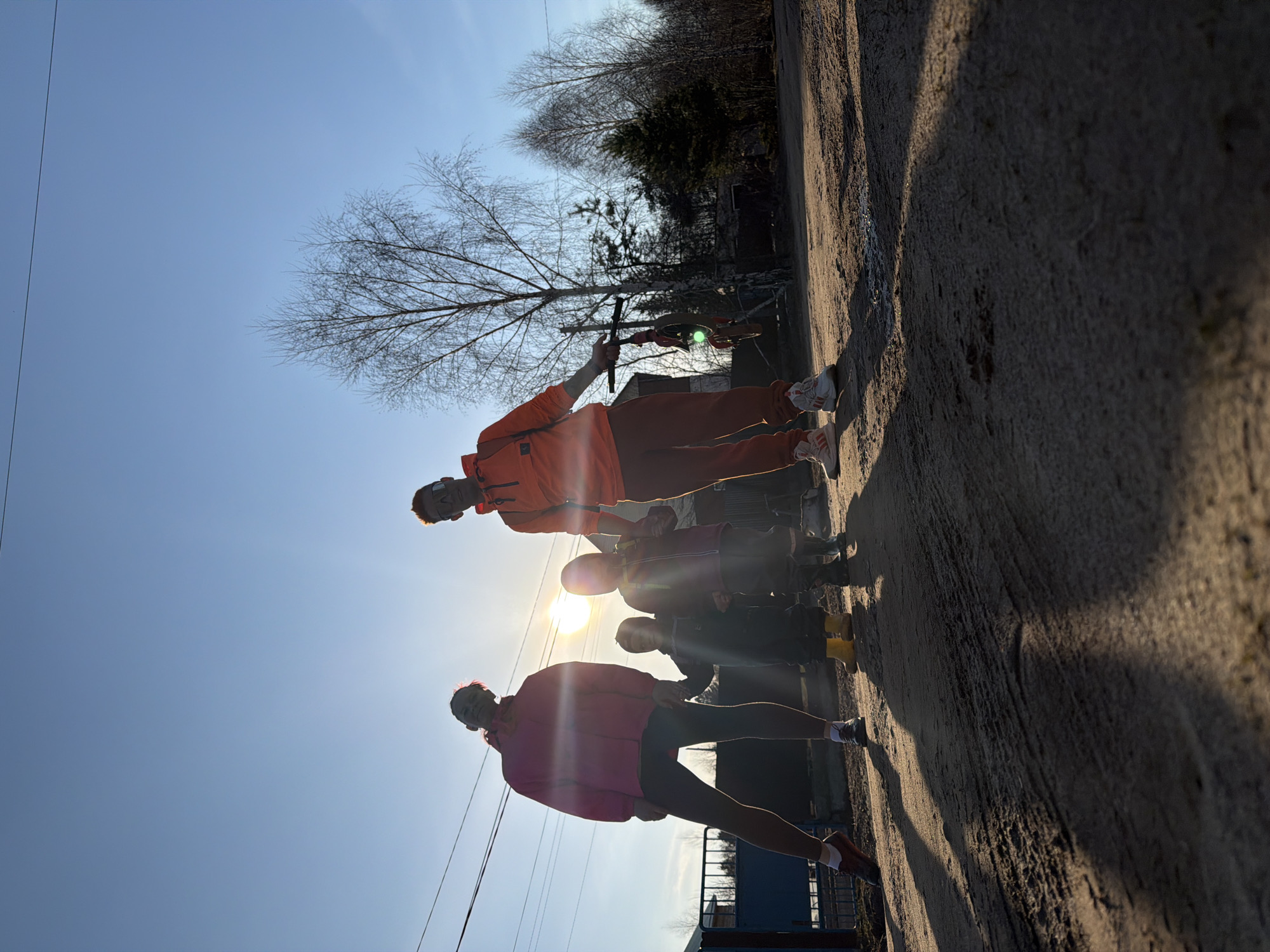 The image shows a group of people standing on a concrete surface outdoors. There are at least 5 people. One person is wearing a pink jacket and black pants, while another person is wearing an orange sweatsuit. The sun is positioned behind the group, creating a silhouetting effect. The sky is blue. A few bare trees are visible in the background, along with a possible building structure. The location is Chaikovsky, Russia.