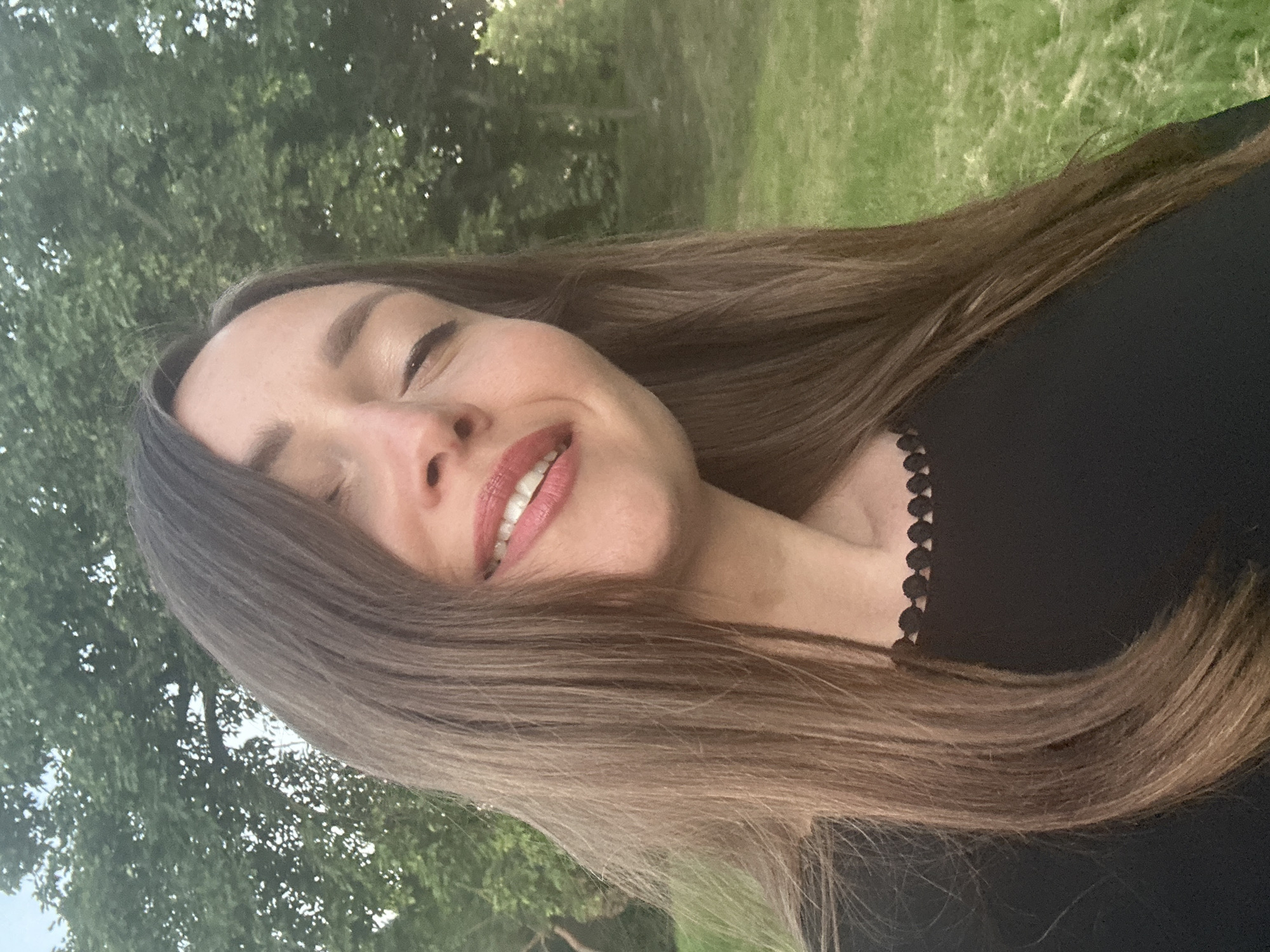 The image is a close-up, outdoor selfie of a young woman with long, brown hair and fair skin. She is wearing a black top with a decorative scalloped trim around the neckline. Her eyes are closed, and she has a slight smile on her lips, which are painted with red lipstick. Her hair is partially covering her face and draped over her shoulder.
The background consists of green grass and trees, suggesting a park or natural setting. The lighting indicates it is likely daytime, possibly late afternoon or early evening due to the soft light.
There are no discernable elements in the image that would suggest a specific location within the city of Bila Tserkva, Ukraine. The scene is generic to many outdoor environments.