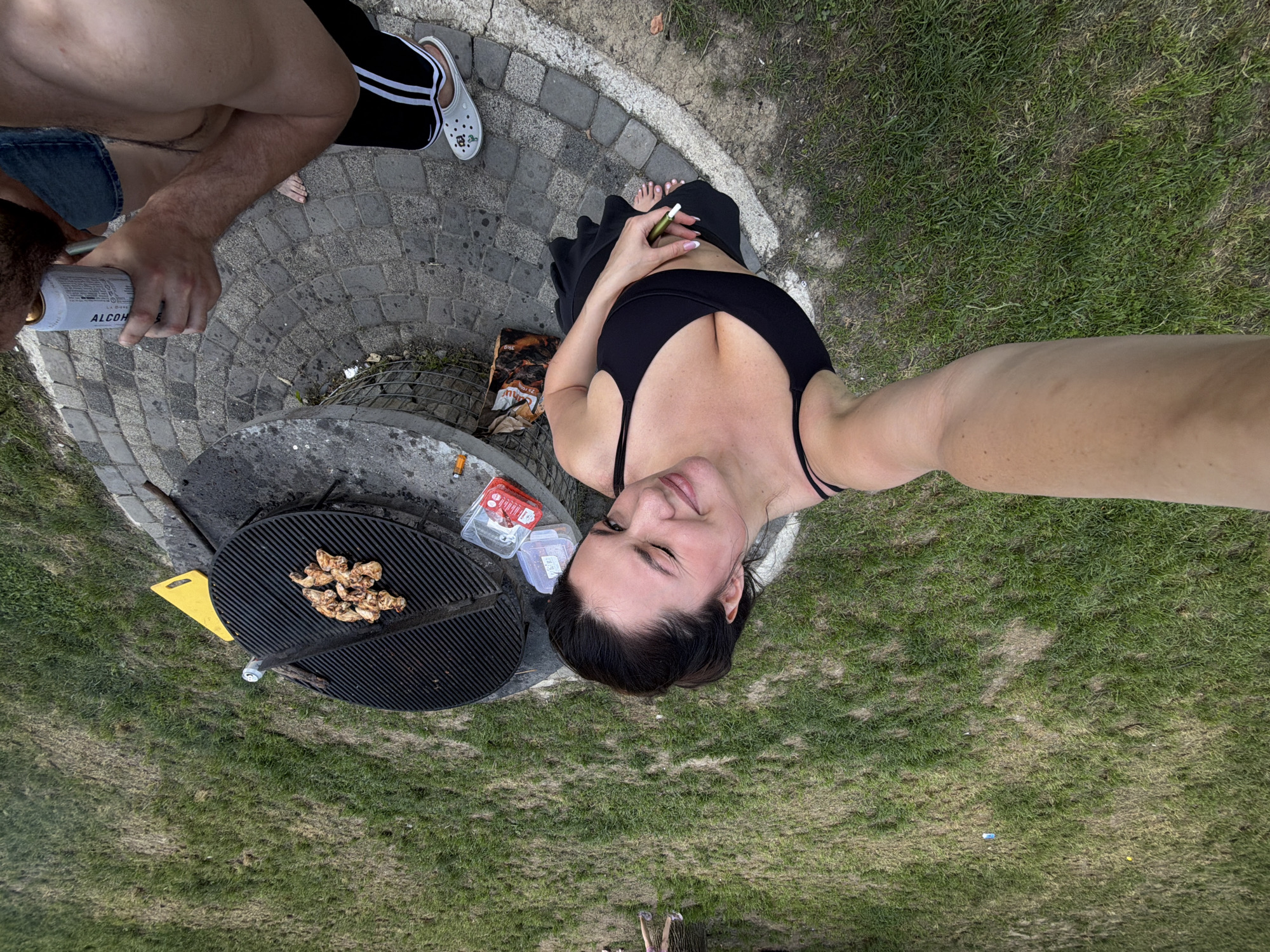 This photograph appears to be taken from a high-angle perspective outdoors, likely at a picnic or barbecue. The location appears to be Cukarica Urban Municipality, Serbia, due to the presence of a Cyrillic “ALCOH” marking on a beverage can.
Several people are visible in the image, though not all fully in the frame. A man holds the aforementioned can. A woman in a black top is the central focus, holding what appears to be a cigarette or similar rolled item. Her body orientation suggests she is taking the photograph.
A grill is visible, sitting on a circular concrete or stone base. There is cooked meat, likely chicken, on the grill. There are several containers and what looks like a bag of charcoal near the grill. The surrounding area is grass. The surface is paved with cobblestones.