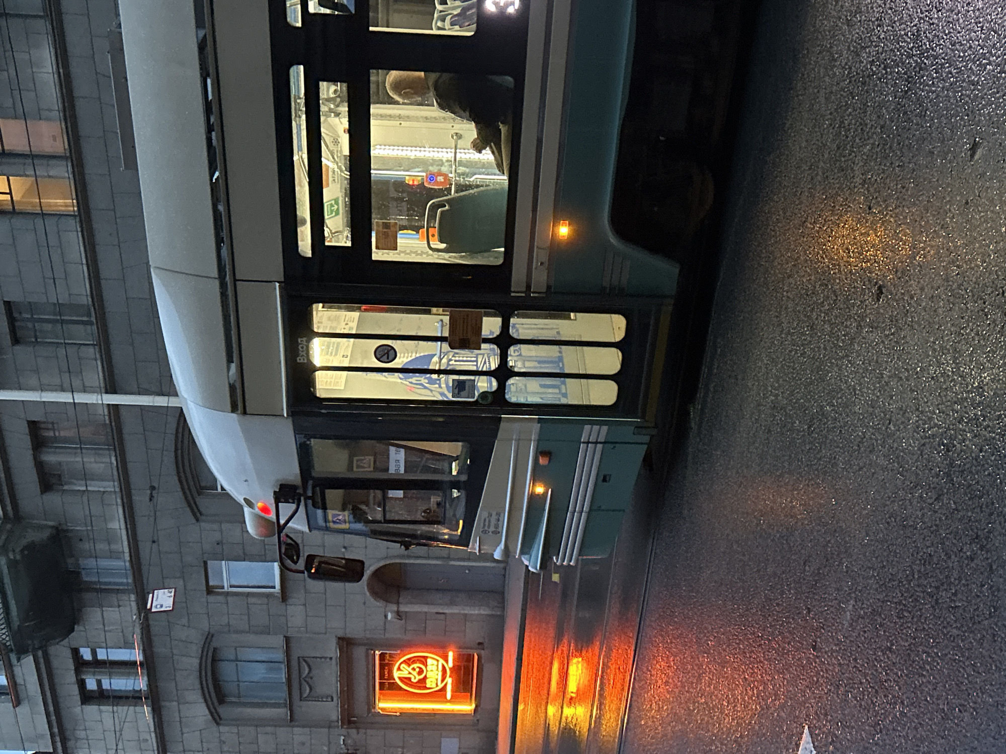 A tram is visible in the foreground, stopped on a wet city street. The tram is teal and white, with its doors open. Inside, a bald man with his back to the camera is seated. The street reflects the orange glow of a neon sign in a storefront to the left of the tram. The storefront has a yellow neon sign with a red circle containing a symbol that appears to be a stylized peace sign. A building with multiple windows stands behind the tram, and overhead wires are visible. The scene appears to be at dusk or night, with artificial lights illuminating the tram and the storefront.