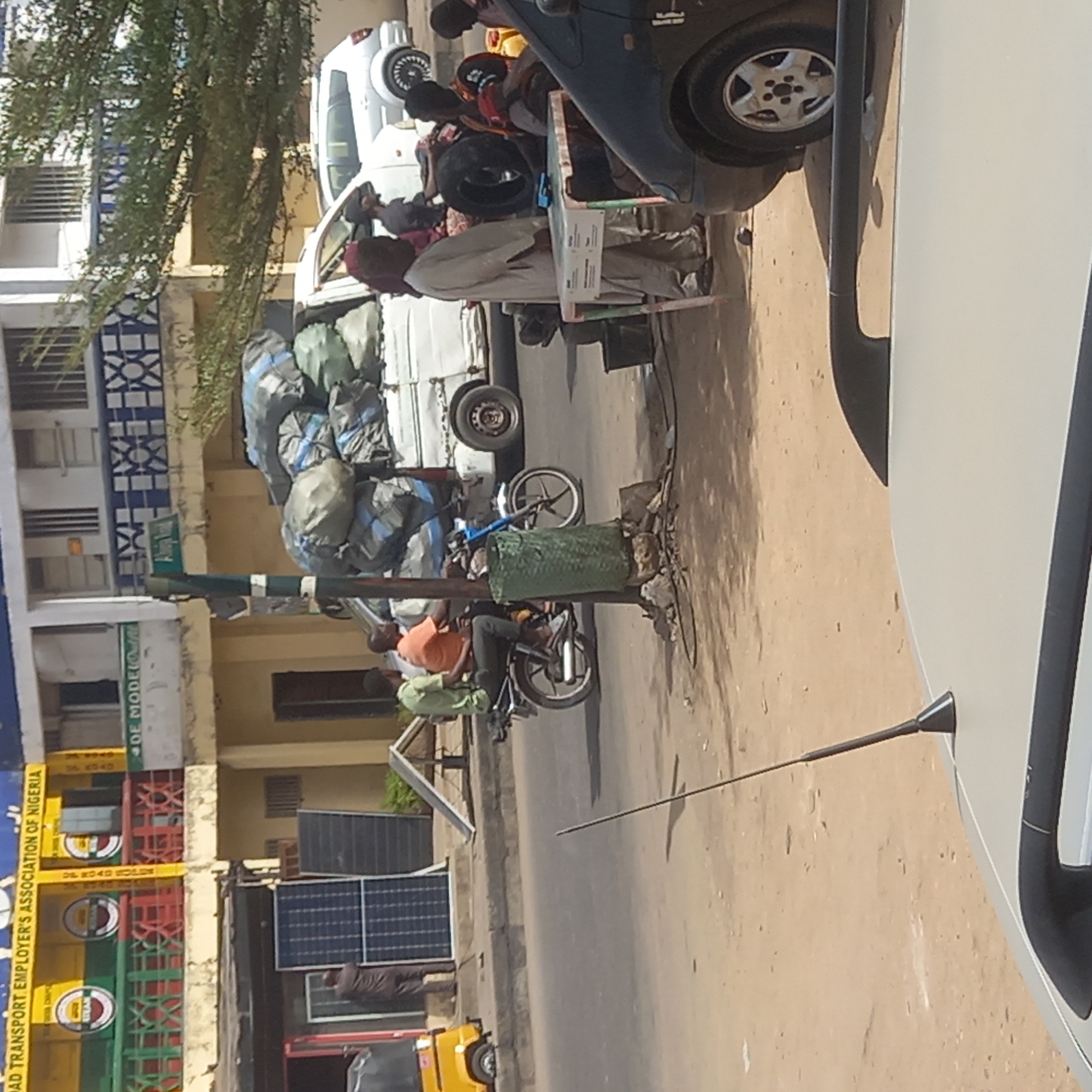The image captures a vibrant, sunlit street scene in Dadi Riba, Nigeria, offering a glimpse into daily life and commerce. The street, likely paved in parts and dusty in others, is bustling with activity.
Several individuals are visible: one person, dressed in a light-colored tunic and a red head covering, stands beside a small, possibly makeshift cart. Nearby, two individuals are seated on motorcycles, one wearing an orange shirt and the other a light green shirt, suggesting they are either waiting or preparing to depart.
The street is populated with various vehicles. A white van or small truck, heavily loaded with numerous large, dark-colored bags (possibly containing goods or waste), is parked on the side of the road. A dark blue car and a white car are also visible, along with multiple motorcycles and the front of a yellow tricycle (keke napep).
Buildings line one side of the street, featuring white and cream-colored facades with horizontal window grates and decorative blue and white patterns. A large, leafy green tree provides shade over part of the scene. Several solar panels are prominently displayed on the ground, leaning against a building, indicating a reliance on alternative energy sources. A tall street pole, partially wrapped in green material, stands in the foreground.
The bright daylight and clear conditions suggest it is midday or early afternoon. The overall atmosphere appears busy and informal, characteristic of a commercial or transport hub.
A notable detail is a colorful sign on one of the buildings, clearly displaying the text: "ROAD TRANSPORT EMPLOYER'S ASSOCIATION OF NIGERIA". Further below, "DR. ROAD ROLE" and "DE MODE" are also partially visible, possibly indicating local businesses or organizations. No strong emotions are discernible from the individuals, who appear to be engaged in their daily routines.
