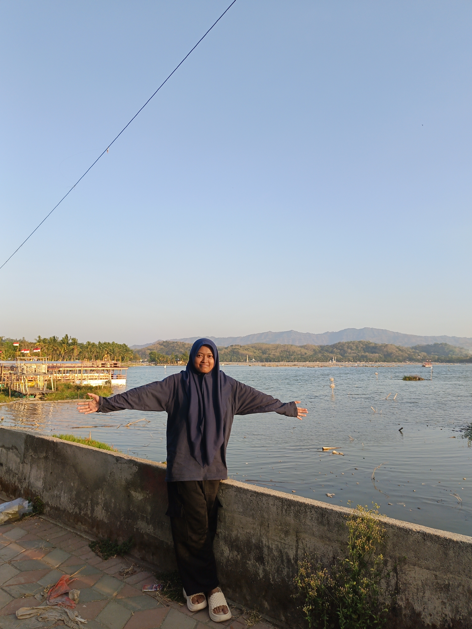 A full shot captures a woman standing in front of a serene body of water, likely in Krakitan, Indonesia, at dusk. She is wearing a navy blue hijab and a loose-fitting navy blue hoodie with dark pants and white sandals. Her arms are outstretched horizontally, palms facing upwards, as if embracing the environment. Behind her, the water is calm, dotted with a few floating objects and poles. In the distance, a fleet of boats, some adorned with Indonesian flags, are docked near the shore, lined by lush green palm trees. The background features rolling hills and mountains under a clear, pale blue sky. The foreground shows a textured concrete wall and a tiled walkway, with some scattered debris and a small patch of green plants.