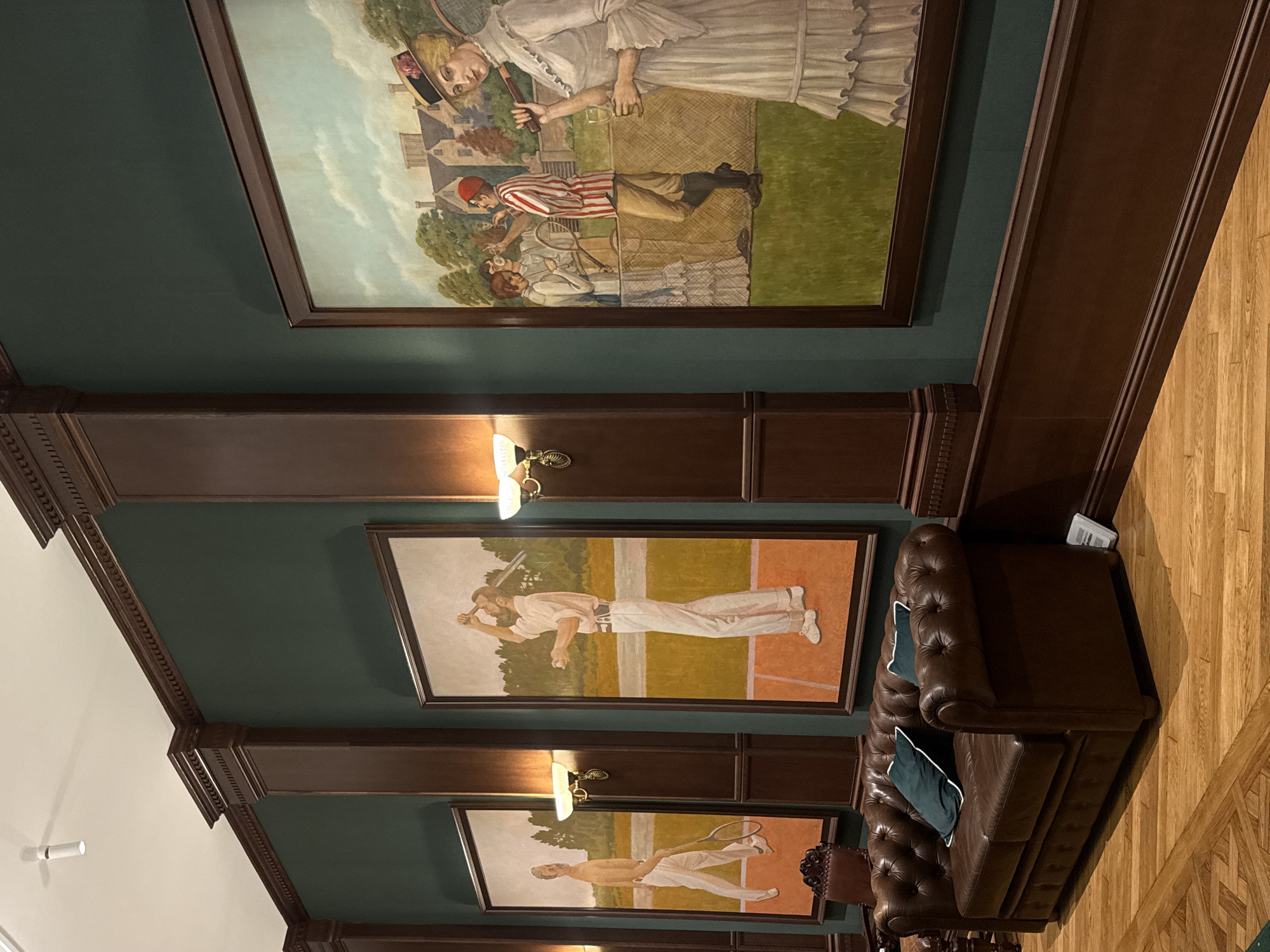 This media file captures a dimly lit interior space, possibly a room within a residential or recreational building. The focus is on several large, framed paintings displayed on a dark green wall, with dark brown wooden paneling and trim framing the artwork and walls.
The topmost painting depicts a scene of people playing a game, possibly tennis, in an outdoor setting with a backdrop of houses and trees. A woman in a white dress holding a tennis racket is prominent in the upper portion of the painting. Below her, figures in period clothing are visible, engaged in some activity on a grassy court.
Below this, a second painting shows a man dressed in a white baseball uniform, holding a bat and a ball, on a baseball field. The setting appears to be a sunny day, suggested by the bright lighting within the painting.
A third painting, partially visible at the bottom, also features a figure in athletic attire, appearing to be holding a tennis racket.
In the foreground and to the right, a dark brown leather Chesterfield sofa with tufted cushions and dark green accent pillows is visible. The sofa is placed on a polished wooden floor with a parquet pattern. The overall ambiance suggests a sophisticated and perhaps historical decorative scheme. The lighting in the room appears to be artificial, coming from wall-mounted sconces positioned above the paintings. There is no visible text in the media file.