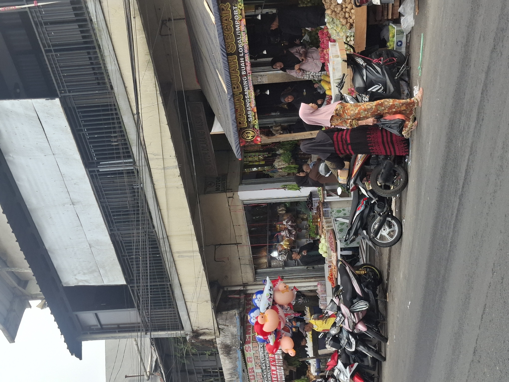 The image depicts a street scene in Gekbrong, Indonesia. A row of shops selling fruits and vegetables line the street. In the foreground, several motorcycles are parked on the asphalt road. A group of people, mostly women in headscarves, are browsing the produce. Some are interacting with vendors, while others are carrying shopping bags. Balloons shaped like animals are displayed outside one of the shops. The buildings appear to be old, with weathered facades and balconies. Overhead, electrical wires are visible. The overall atmosphere suggests a busy local market.