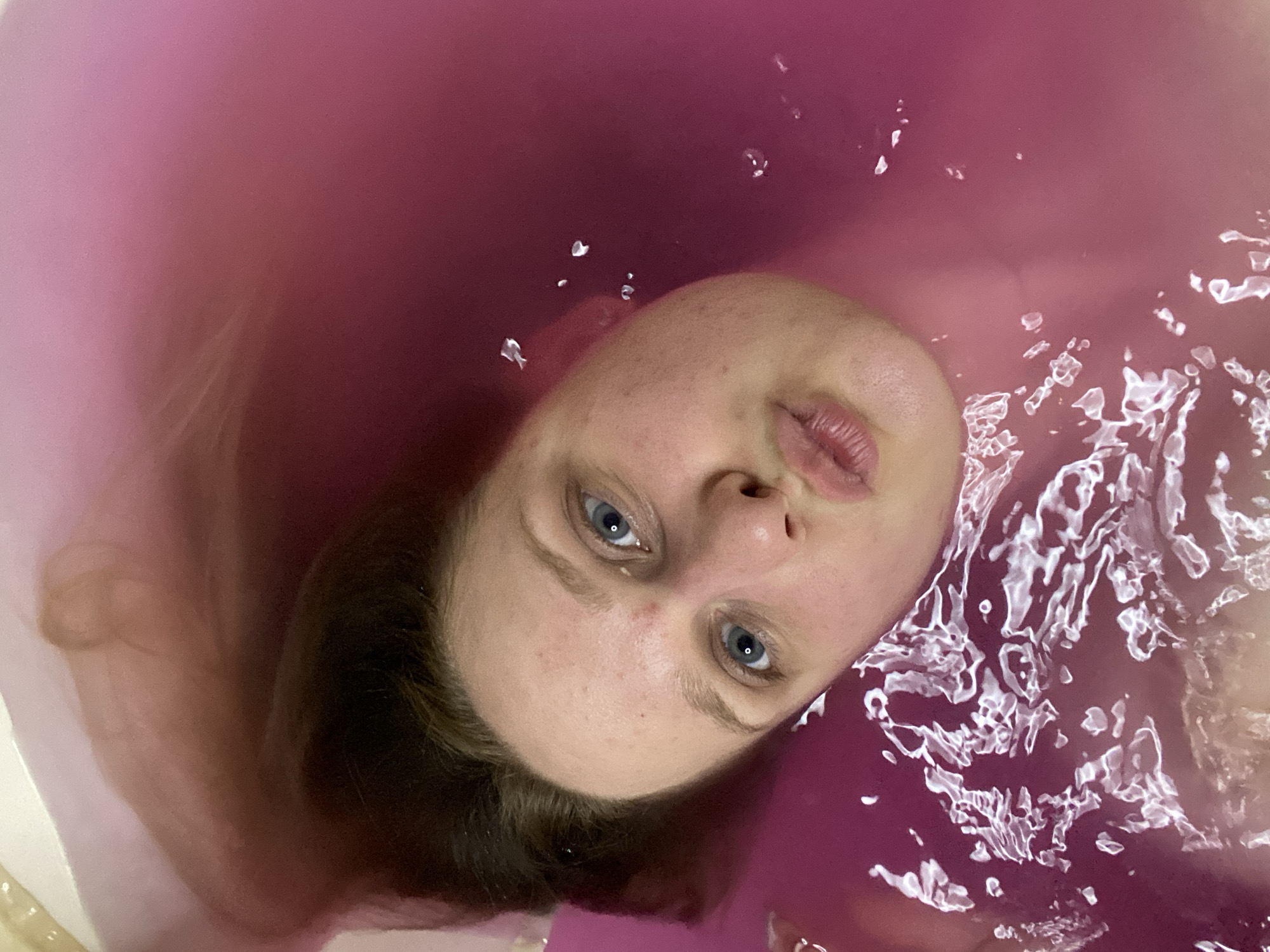 An overhead, close-up shot captures a person's face submerged in pink-colored water, likely a bath. The individual's head is tilted, with their face oriented towards the camera. They have light-colored skin and blue eyes, and their lips are pursed. Their dark brown hair is spread out in the water around their head. The water is disturbed in some areas, creating ripples and reflections. The context of Chaikovsky, Russia, is not discernible from the visual information present in the image.
