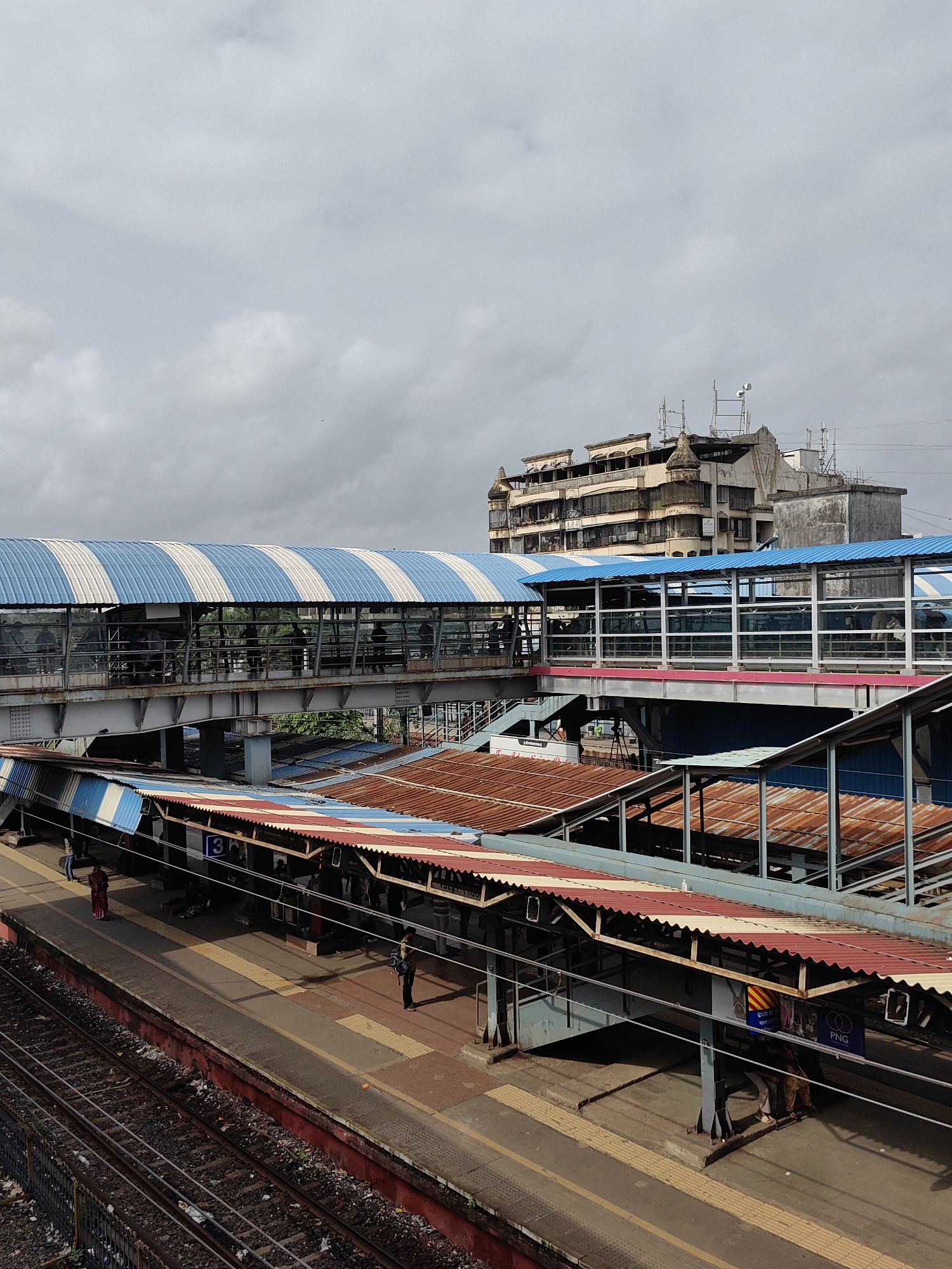 The image depicts an elevated pedestrian walkway at a train station in Mira-Bhayander, India. The walkway, with its blue and white striped roof, spans across multiple train platforms. Below the walkway are platforms covered with red and white striped roofs, some appearing weathered and rusted. Several individuals are visible on the platforms and within the walkway. Train tracks run in the foreground. In the background, an older, multi-story building with a distinctive architectural style stands against a cloudy sky. The scene is an outdoor, public transportation hub under overcast weather conditions.