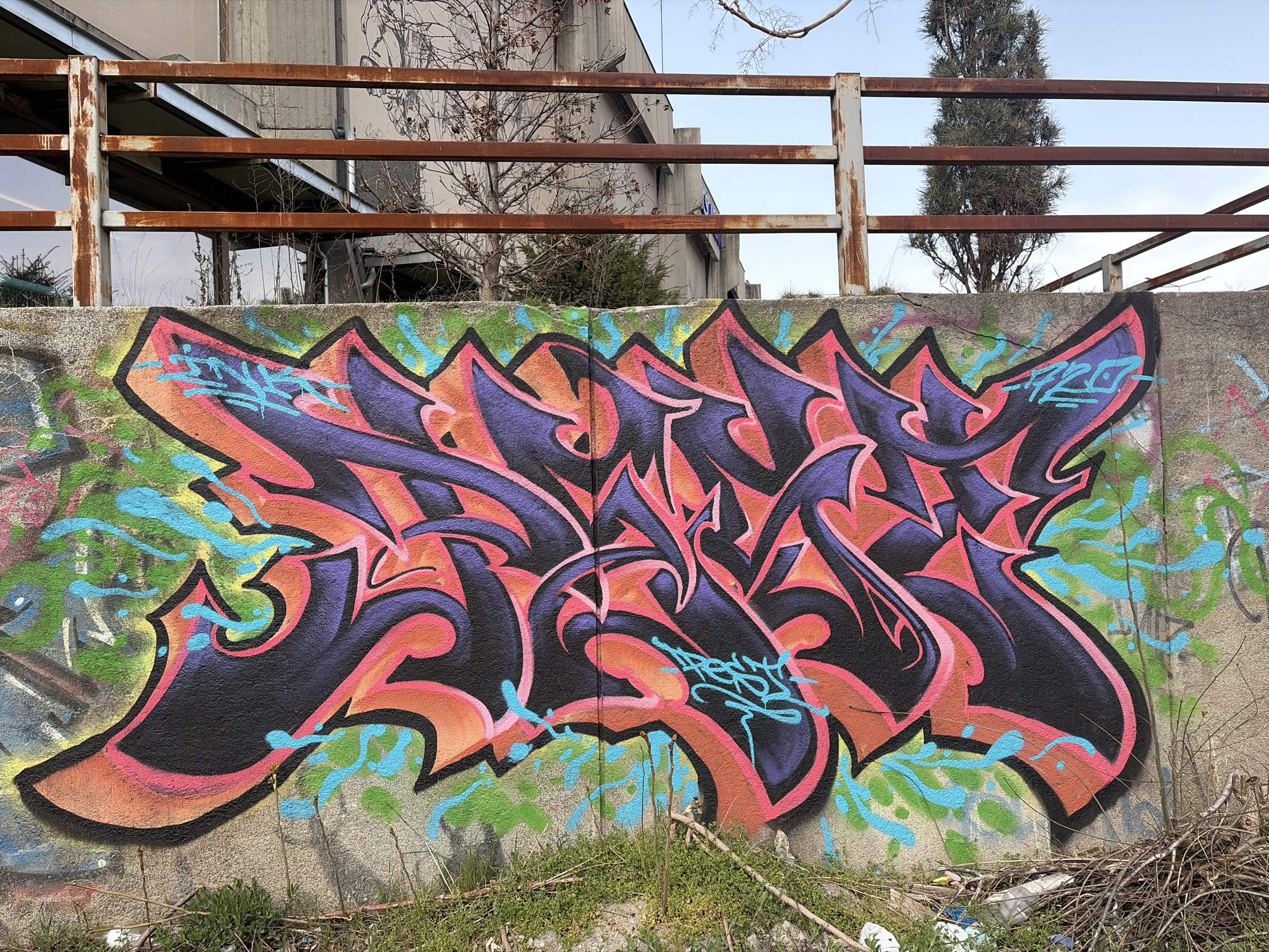 A concrete wall features a large, intricate graffiti artwork in shades of black, purple, orange, and pink. The artwork is accented with blue highlights and lettering. The wall is also marked with green and blue paint splatters. Tufts of grass and small branches are growing at the base of the wall. Behind the wall, a rusty metal railing runs horizontally. Beyond the railing, a multi-story building with visible windows and a tree are visible. The sky above is pale blue. The location is most likely in a desolate area of Sofia, Bulgaria, judging by the graffiti and the architectural style.