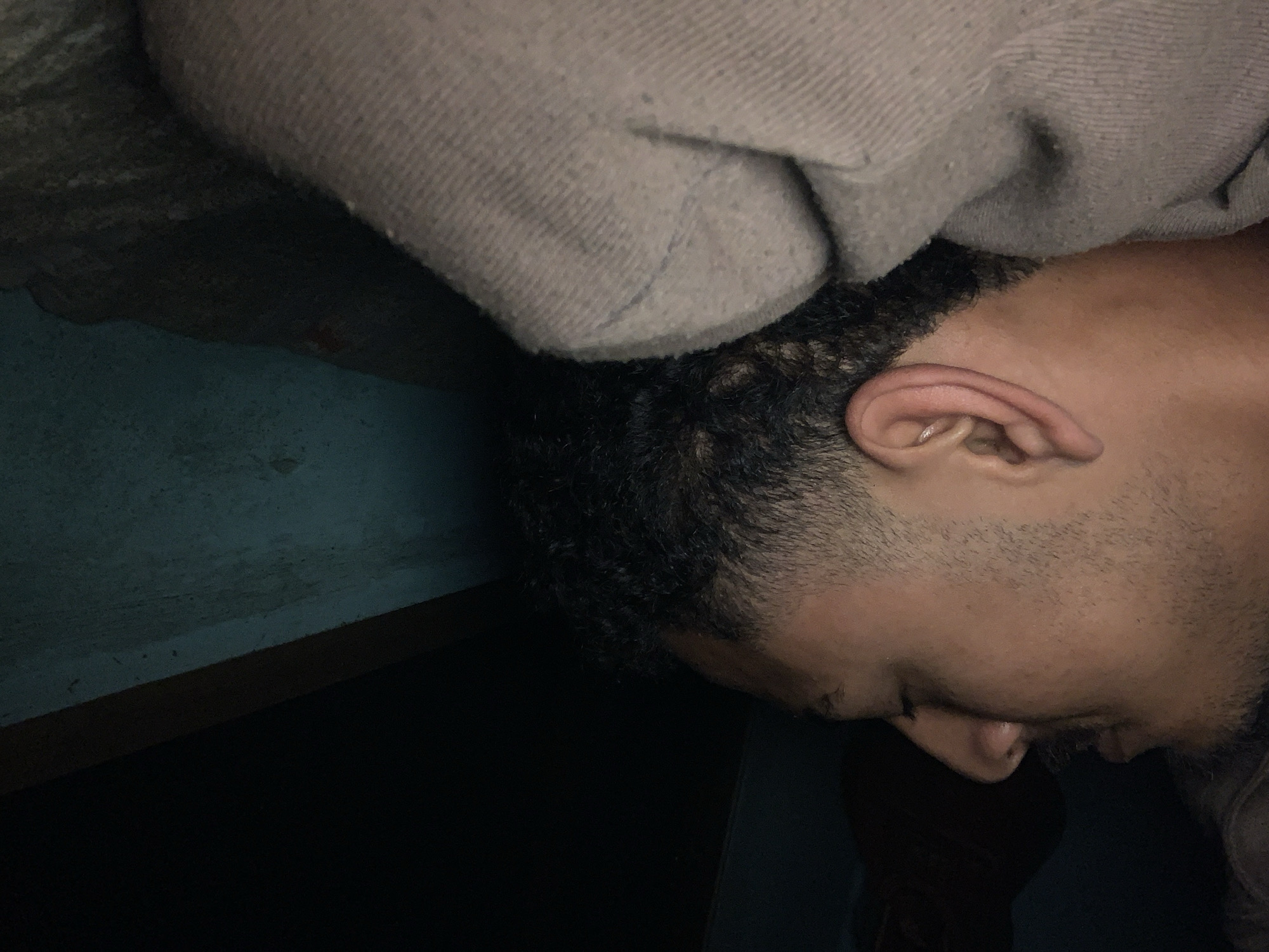 A close-up, profile shot of a man sleeping, face turned away from the camera, is captured in Santa Ana, Venezuela. The man, lying on his left side, is covered by a light gray, textured blanket that obscures his body from the chest down. His right ear, prominently visible, is a pale pinkish hue. He has dark, curly hair and a short, stubbly beard. The background is dark, with a hint of a dark blue wall and a wooden surface visible to the right. The lighting is dim, suggesting an indoor, possibly bedroom setting, with shadows obscuring many details.