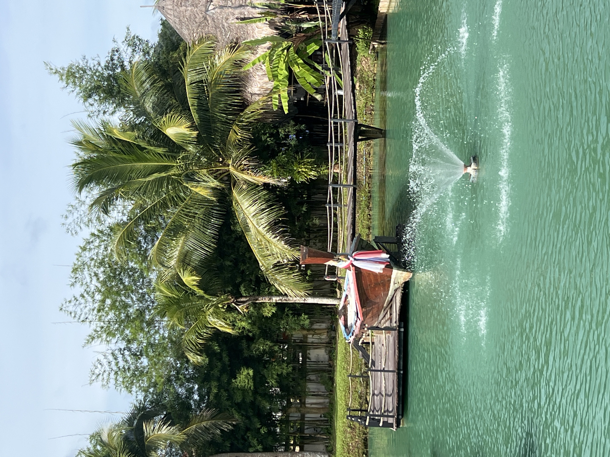 The image depicts a serene tropical scene in Tambon Pa Kae Bo Hin, Thailand. A traditional Thai longtail boat, painted red with blue accents, is moored to a wooden pier. Colorful ribbons are tied to the boat's prow. In the foreground, a fountain sprays water in the vibrant turquoise water. In the background, lush green palm trees, bamboo stalks, and a thatched-roof structure create a dense tropical environment. A wooden bridge extends across the water in the mid-ground. The sky is clear blue with scattered white clouds. No people are visible in the image.