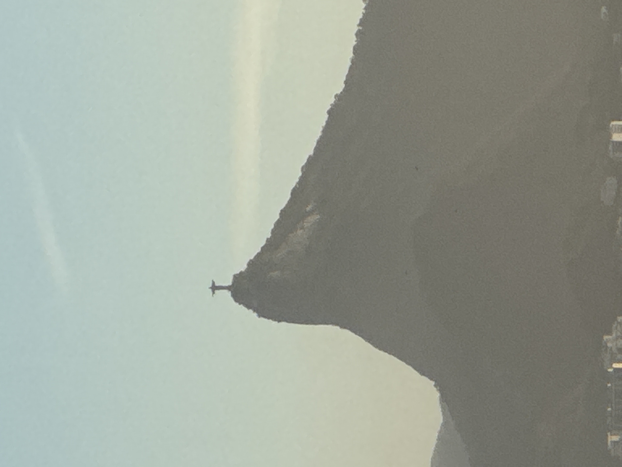 The image displays a wide shot of Christ the Redeemer statue perched atop Corcovado Mountain, a prominent landmark in Rio de Janeiro, Brazil. The statue is silhouetted against a hazy sky, with the mountain itself appearing as a dark, imposing mass. The lower portion of the image reveals the distant cityscape of Niterói, characterized by indistinct buildings and structures that fade into the atmospheric haze. The overall tone of the image is muted, with a soft light and a limited color palette dominated by shades of gray and pale blue, suggesting either dawn or dusk.