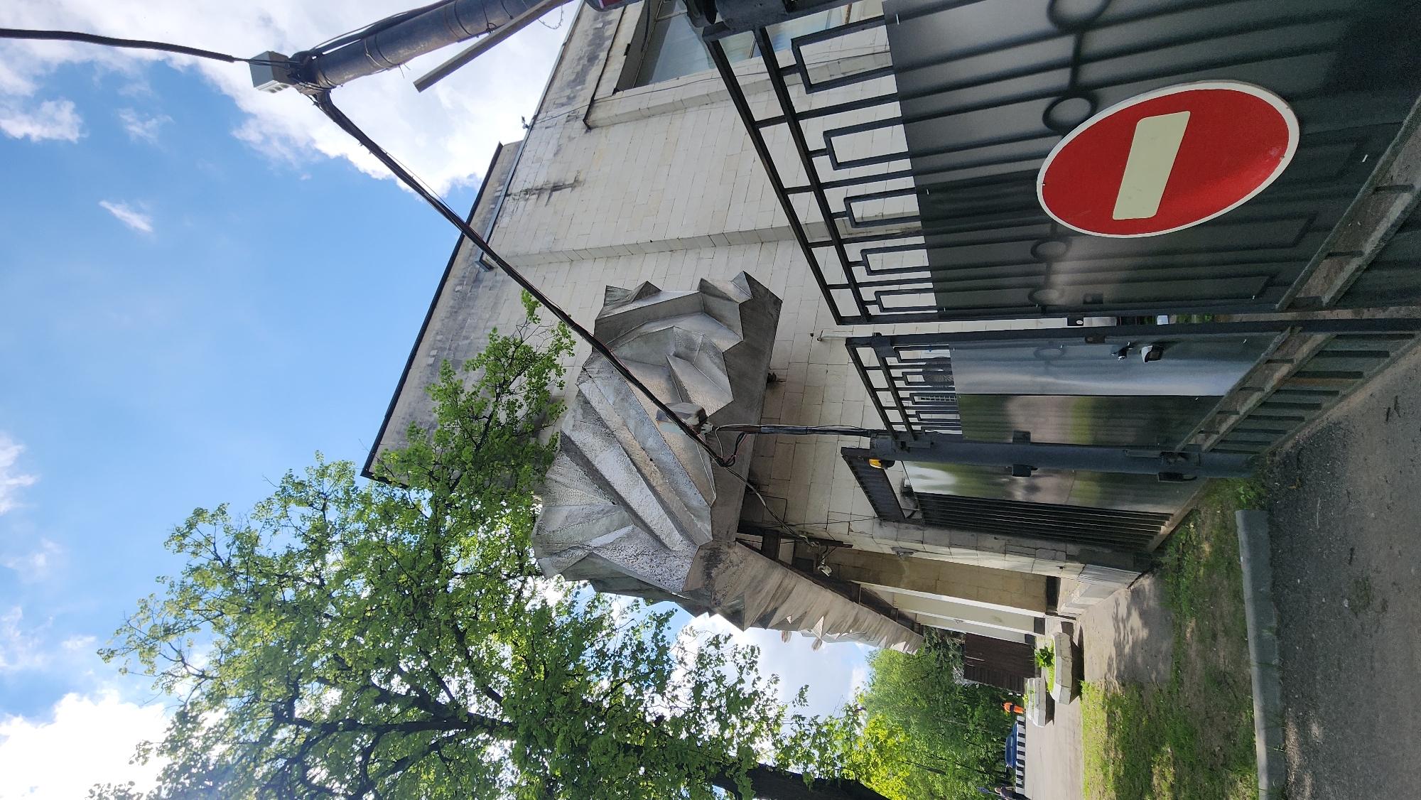 The image shows a building with a distinctive, geometric, silver-colored sculptural element attached to its facade. A tree with green leaves partially obscures the view of the building and sculpture. A metal fence with a red and white "No Entry" sign is visible in the foreground. The image was taken in Khimki, Russia.