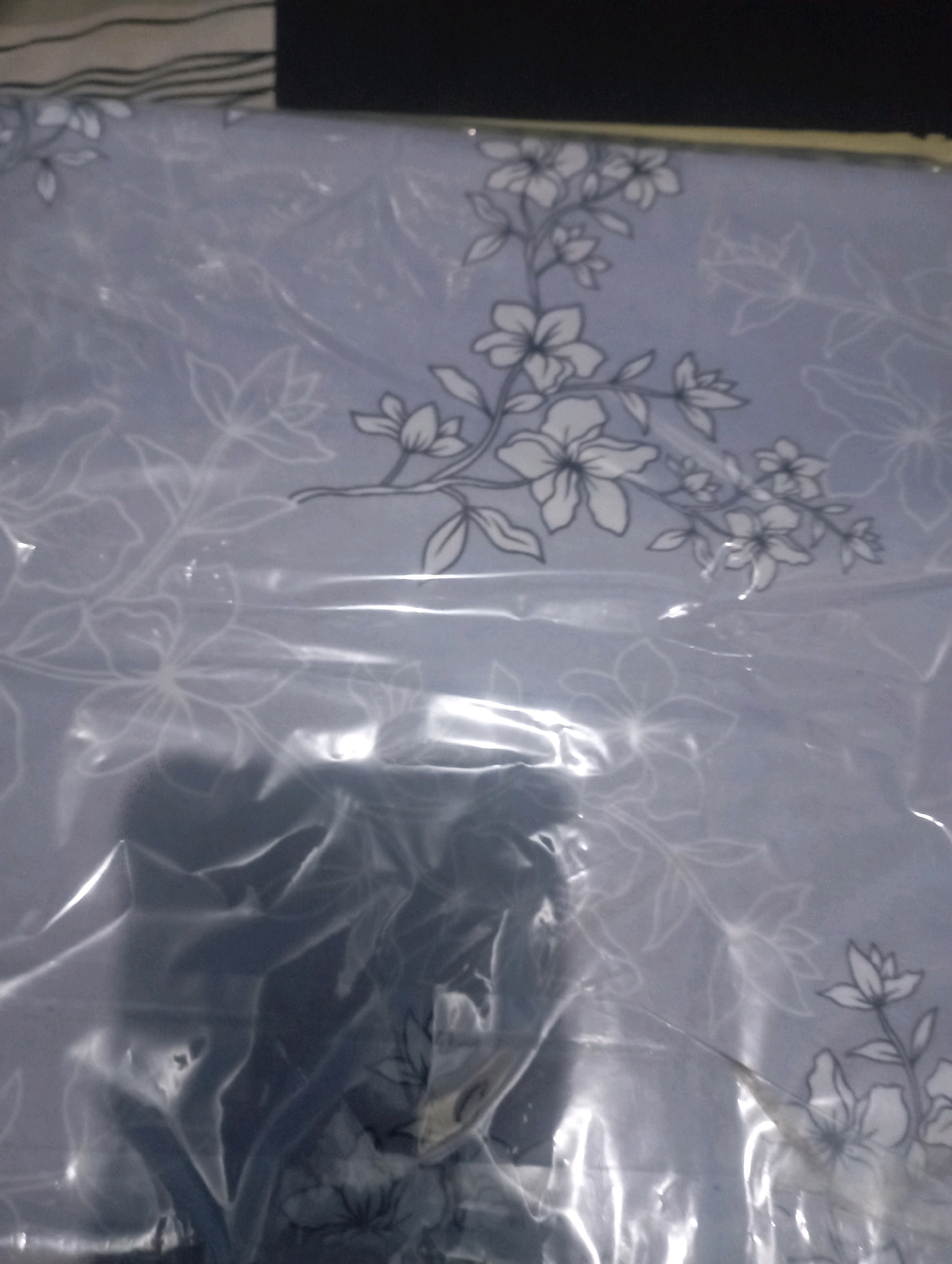 The image depicts a close-up, slightly blurry view of a light purple fabric with a delicate floral pattern. The fabric is covered in a transparent plastic wrap, which creates reflections and obscures some details. The floral design consists of outlines of flowers and leaves in a darker shade, possibly gray or a muted blue. The flowers are depicted with multiple petals, and the leaves are elongated and pointed. Some parts of the fabric are more clearly visible, showing a cluster of flowers and branches, while other areas are covered by the plastic's glare and folds. The background of the image shows horizontal stripes in dark and light shades at the top, suggesting the presence of bedding or another textile. The overall impression is of a new textile product, possibly a bedsheet or fabric intended for home furnishings, still in its packaging. There are no discernible elements to indicate its location within Senangsari, Indonesia, or any specific environmental context.